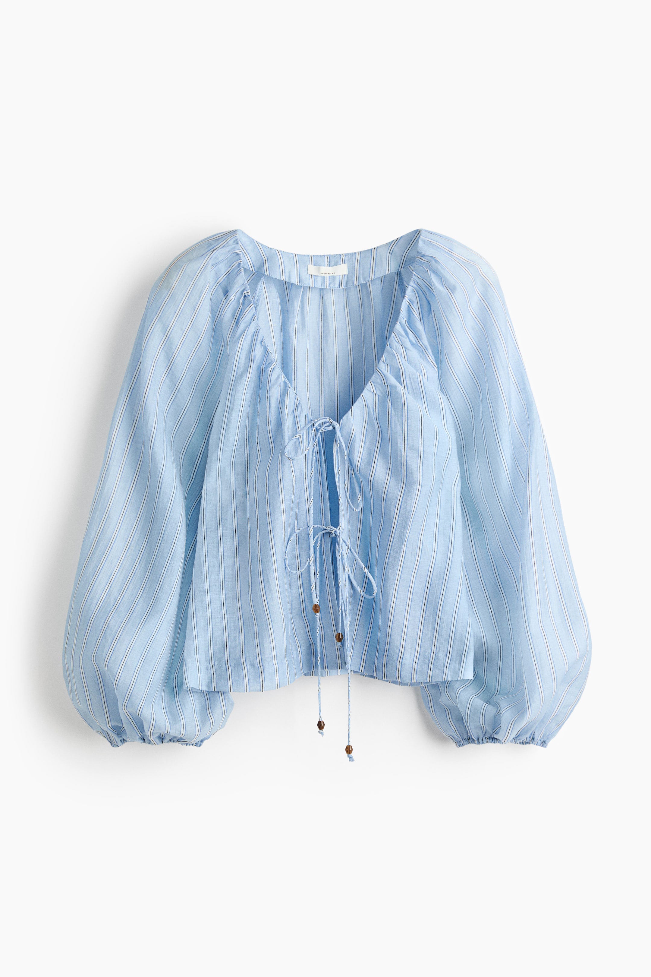 Ver imagem maior: Uma blusa azul-clara com finas riscas verticais brancas e azul-escuro apresenta um corte descontraído e curto. Foi desenhada com um decote redondo e aberto, onde duas tiras finas com pequenas contas de madeira castanha nas extremidades se cruzam e atam no centro da frente. As mangas compridas são volumosas e reúnem-se em punhos elásticos.