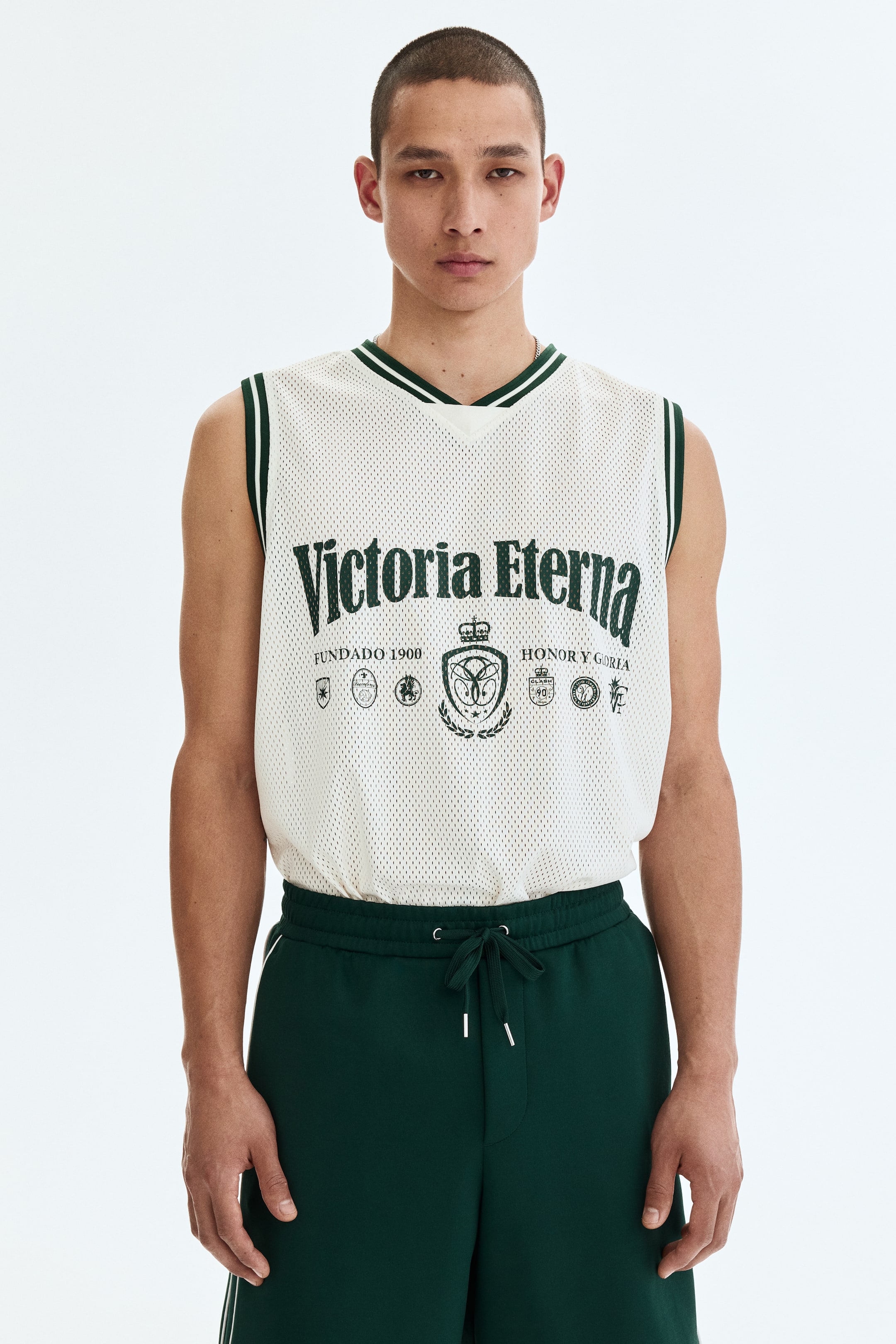 View larger image: An off-white mesh jersey top featuring a V-neck and armholes with green and white striped trim, adorned with a large dark green 'Victoria Eterna' graphic and crests on the chest. Dark green sweatshorts complement the top, featuring a drawstring waist with silver aglets and white side stripes.