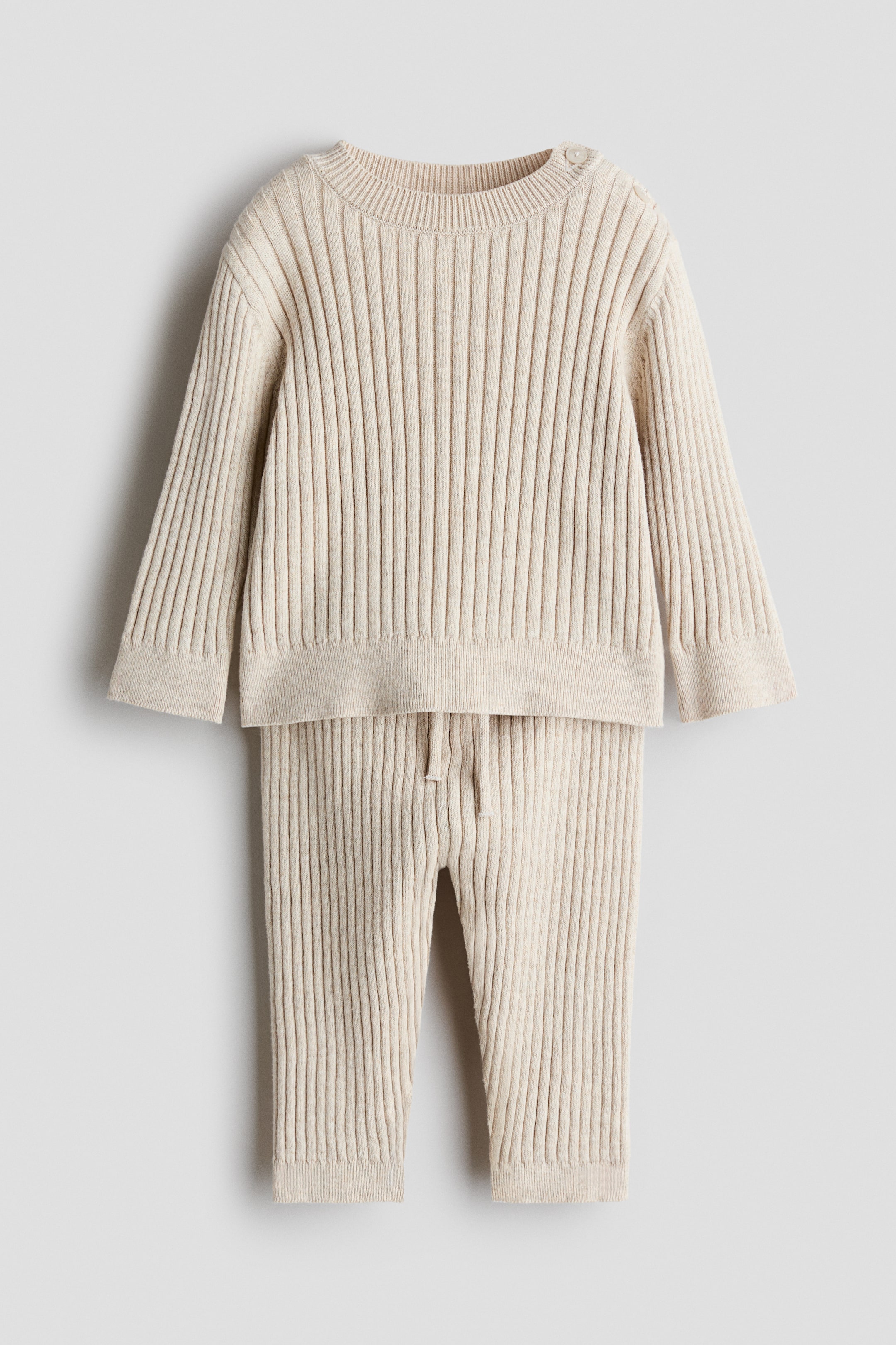 Ver imagen más grande: Un conjunto de punto de algodón beige de canalé ancho, compuesto por un jersey y un pantalón, se muestra de frente. El jersey presenta botones en un hombro y acabados de canalé fino en el cuello, los puños y el bajo, mientras que el pantalón muestra una cintura elástica con cordón ajustable y bajos de canalé fino.