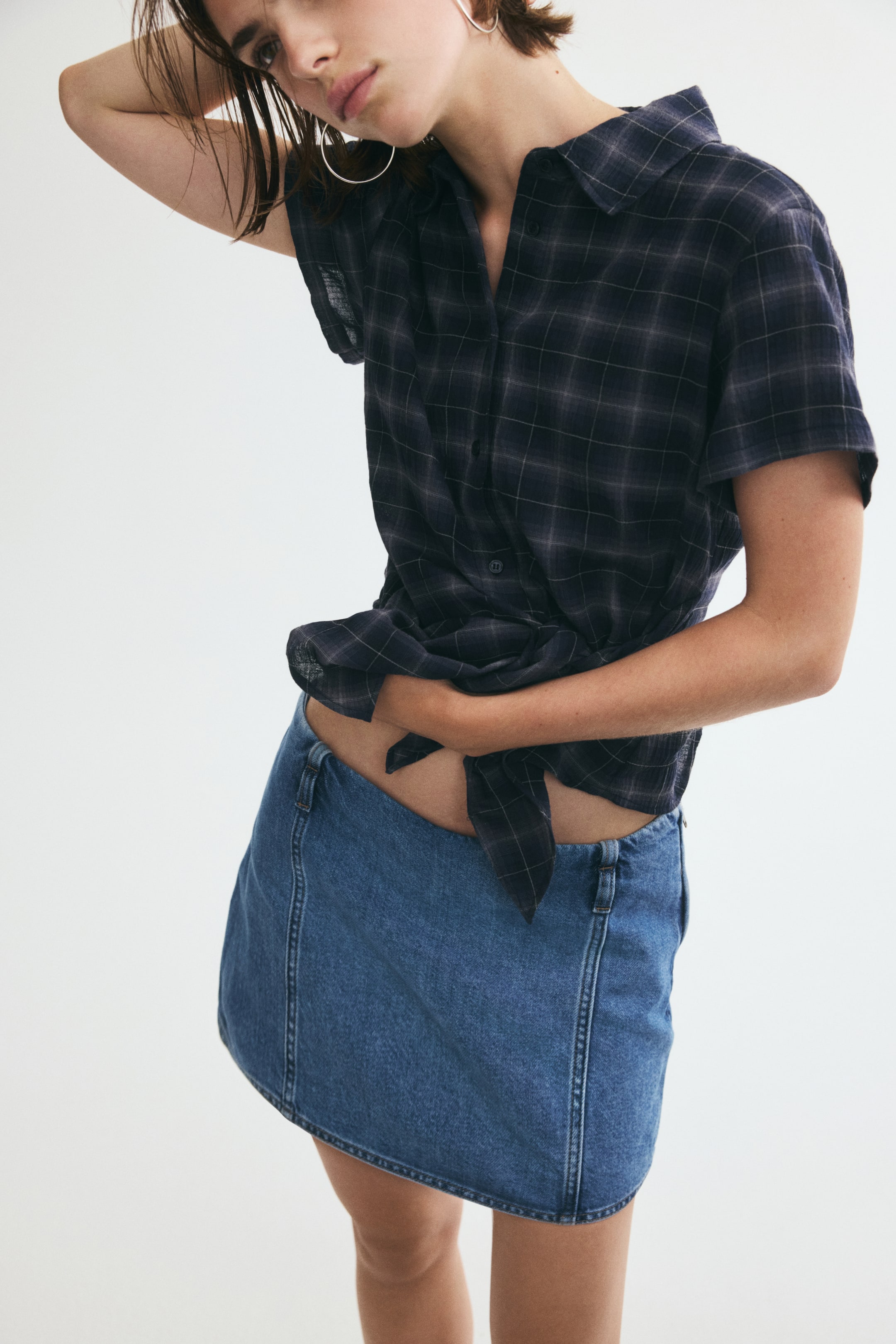 View larger image: A short denim skort in a medium blue wash is worn by the model, featuring vertical seam details on the front panel and a low-rise waist. The skort is styled with a dark, short-sleeved plaid shirt tied at the waist.