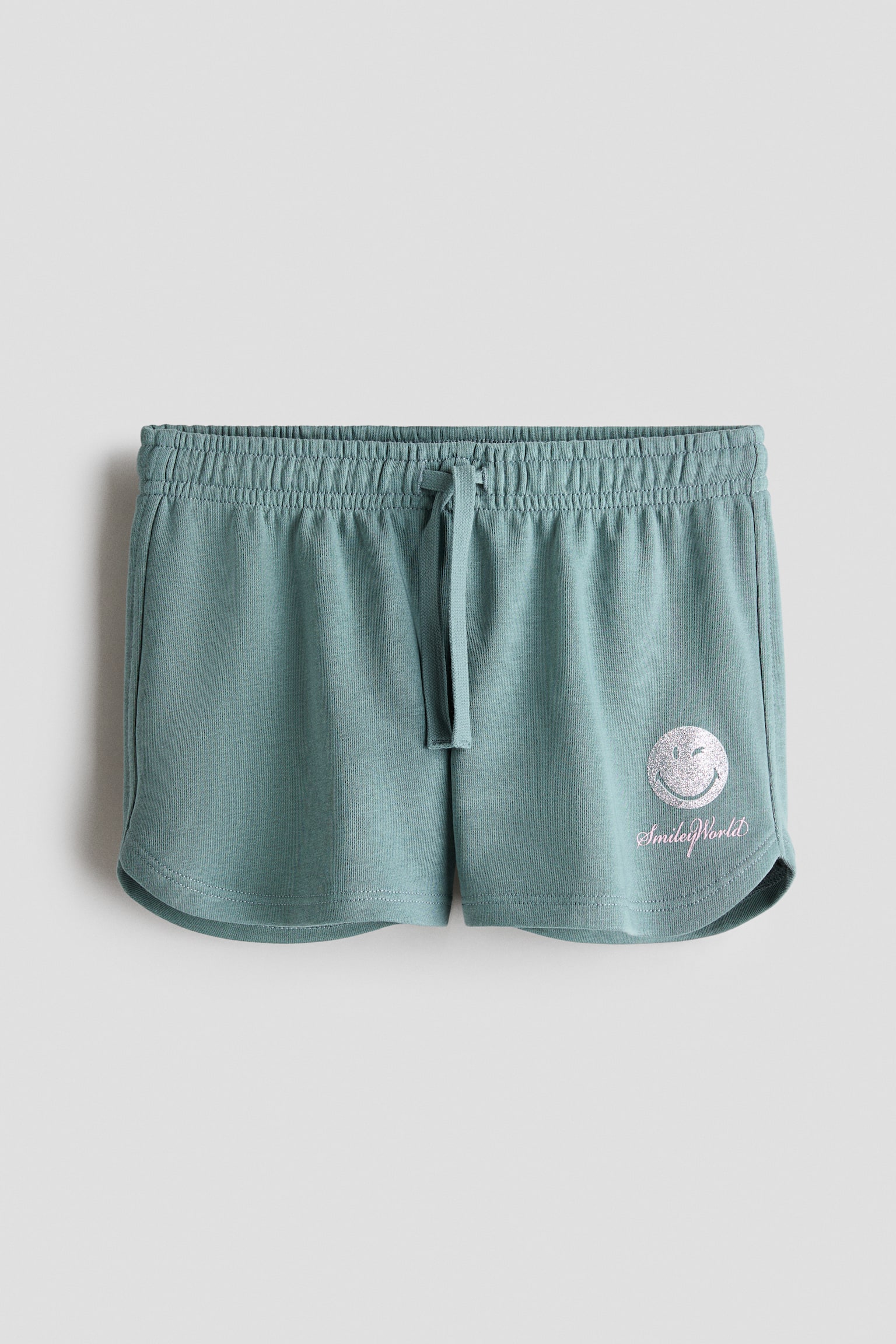 These sage green sweat shorts are shown from the front, featuring an elasticated waistband with a matching drawstring and rounded hems. The left leg displays a silver glittery winking smiley face graphic with pink script text reading "SmileyWorld" underneath.
