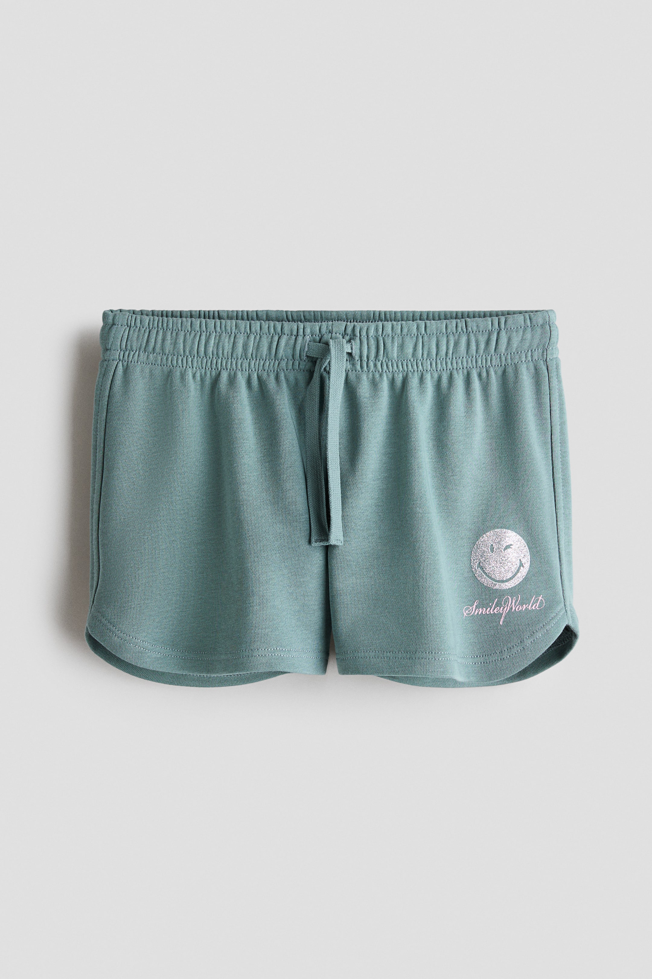 View larger image: These sage green sweat shorts are shown from the front, featuring an elasticated waistband with a matching drawstring and rounded hems. The left leg displays a silver glittery winking smiley face graphic with pink script text reading "SmileyWorld" underneath.