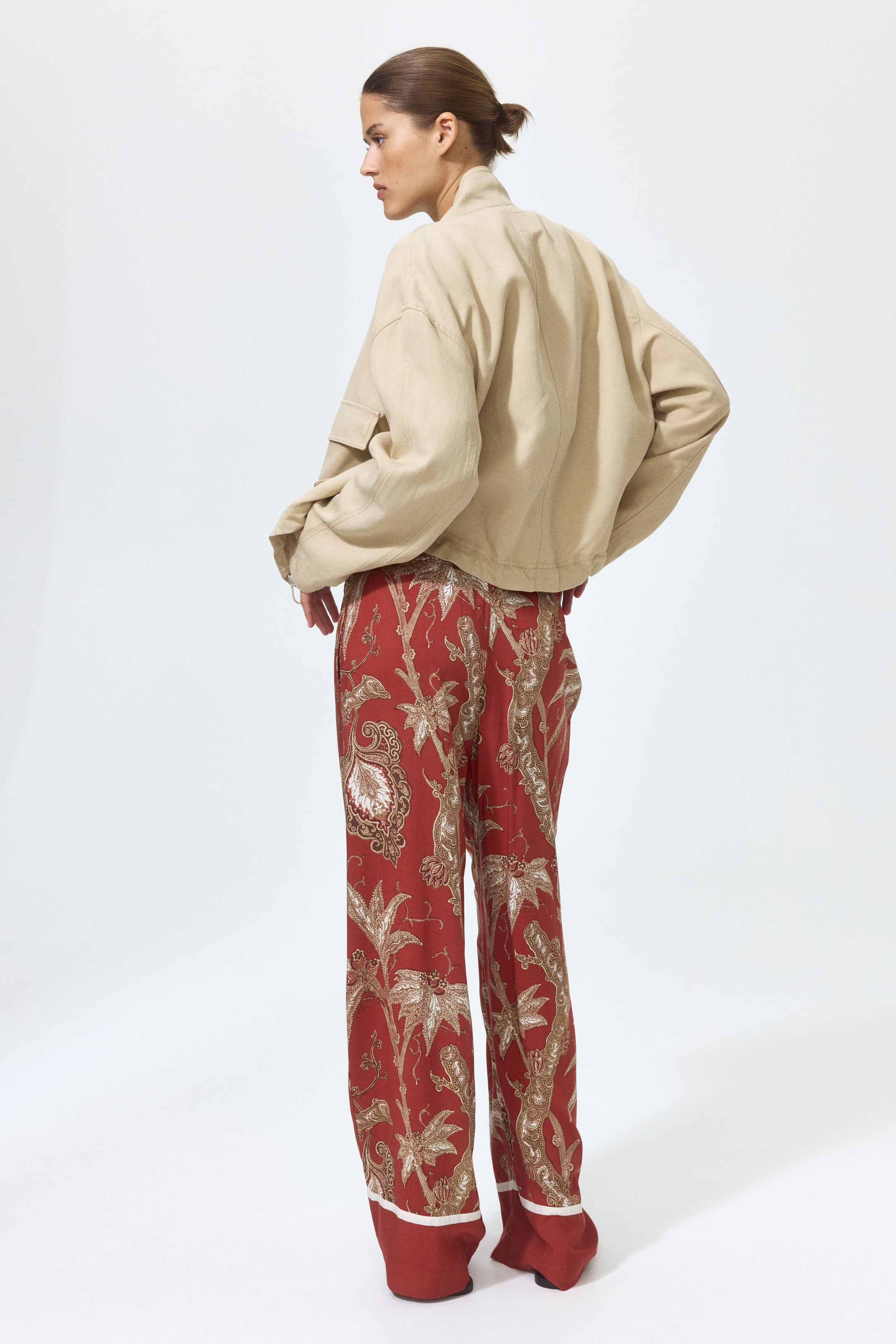 Voir une image plus grande: Pantalon en viscose rouge avec un motif botanique léger, doté d'une taille haute et de jambes larges et droites, porté par le mannequin. Le pantalon présente également une bande blanche à l'ourlet. Le mannequin se tient debout, les mains à la taille, stylisant le pantalon avec une veste beige.