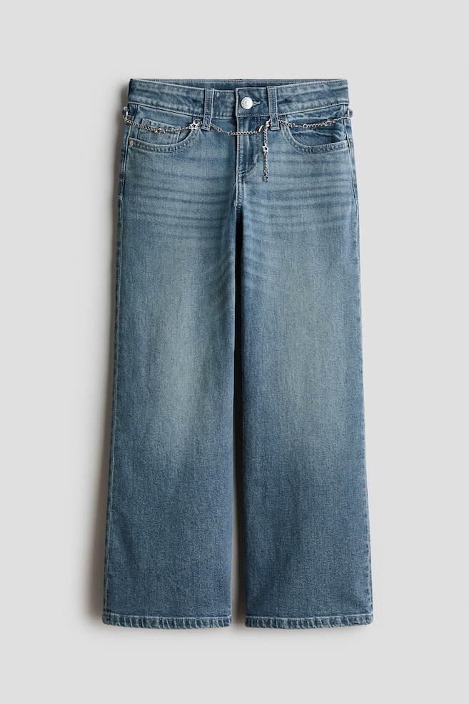 A front view of wide-leg, washed blue denim jeans featuring a low waist with a button fastening and a zip fly. The jeans display a baggy fit with subtle fading on the thighs and are styled with a decorative silver-tone chain with star charms looped through the front belt loops. - 1