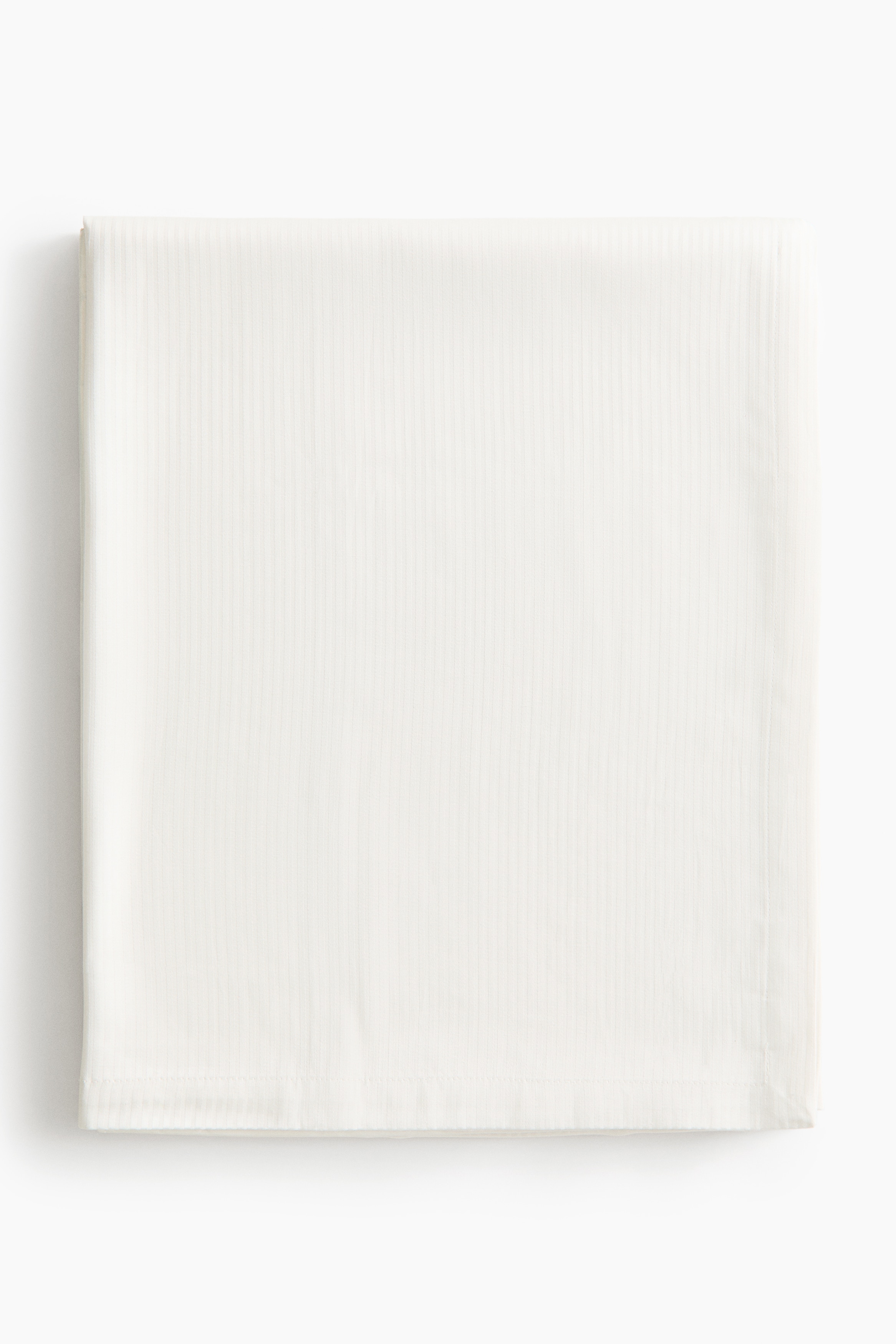 A light beige cotton-blend tablecloth is draping over a rectangular table, showcasing a subtle, matching-tone vertical striped woven pattern. The fabric is hanging with soft folds at the corner, highlighting its textured weave and a wide folded hem along the bottom edge.