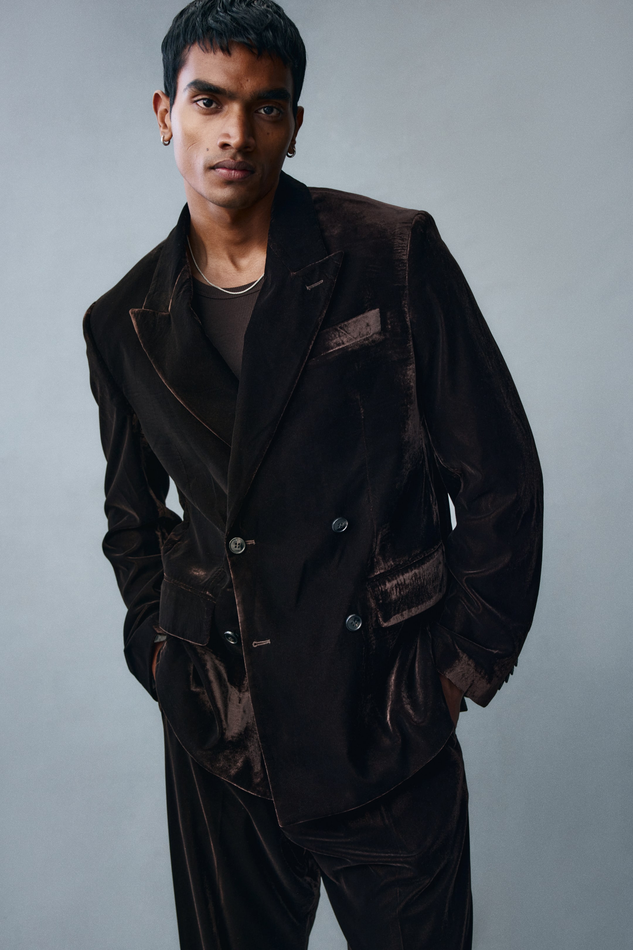 View larger image: A dark brown velvet double-breasted jacket is being worn by the model, featuring peak lapels, front button closures, a chest pocket, and flapped side pockets. The jacket is styled over a brown crew-neck top and paired with matching velvet trousers.