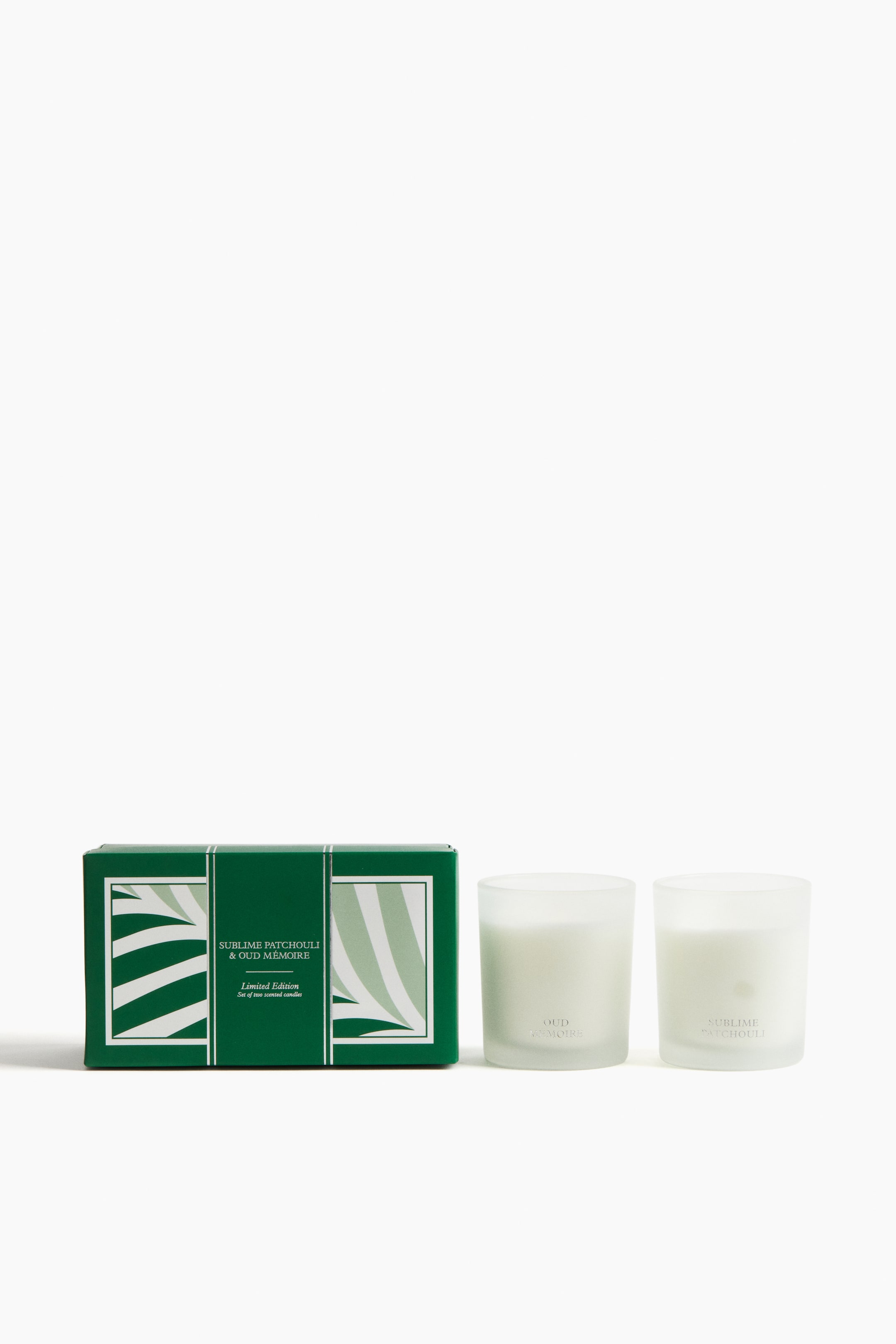 View larger image: A dark green rectangular gift box is facing FRONT, featuring a light green and white striped pattern. Next to the box, two cylindrical frosted glass candle holders are standing upright, each containing white wax and displaying scent names on their FRONT surfaces.