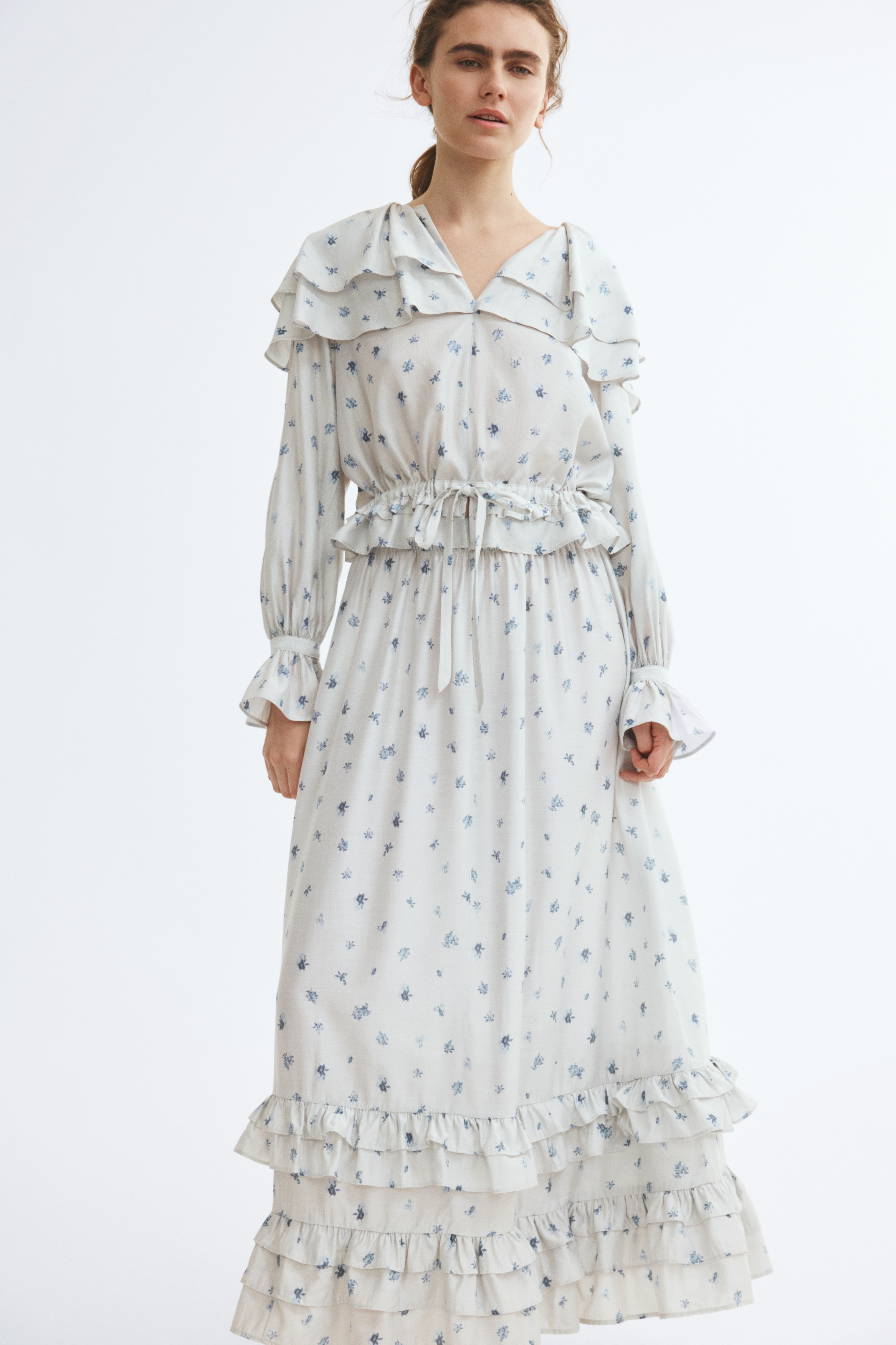 A calf-length, slightly A-line skirt in off-white crinkled chiffon is shown from the front, featuring an all-over blue floral pattern and an elasticated waistband. The lower half shows two tiers of double-layered, frill-trimmed ruffles above the hem.