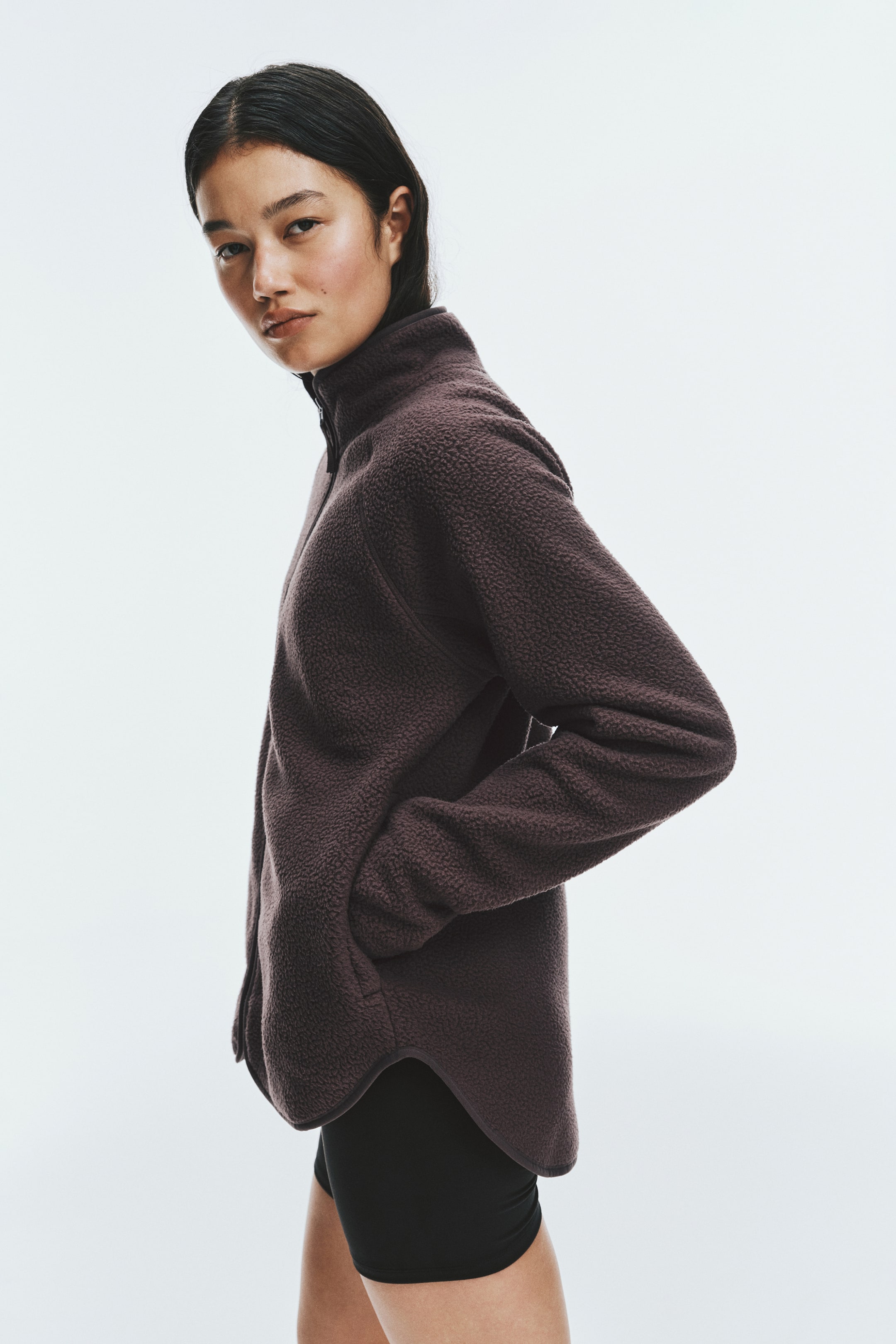 Büyük resmi görüntüle: Model, koyu kahverengi teddy fleece activewear ceket giymiş; yüksek dik yaka, fermuarlı ön tasarım ve arkada daha uzun olan kavisli etek ucu dikkat çekiyor. Model, profilden duruyor ve bir elini fermuarlı yan cebe sokmuş; ceketi siyah atletik şortla kombinliyor.