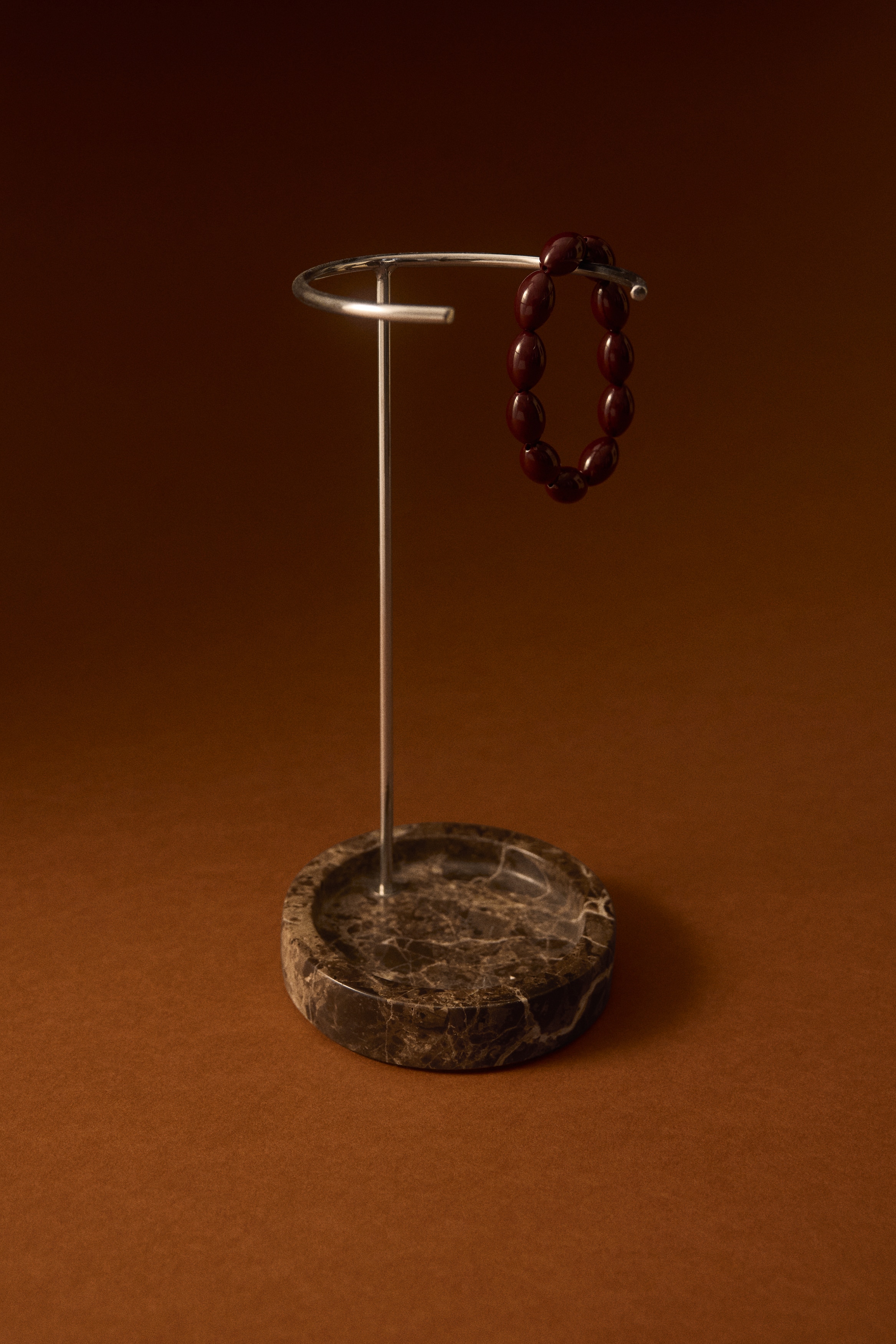 Un espositore presenta una base rotonda in marmo marrone con venature bianche, da cui si erge un'asta metallica color argento che sostiene un anello metallico circolare nella parte superiore.