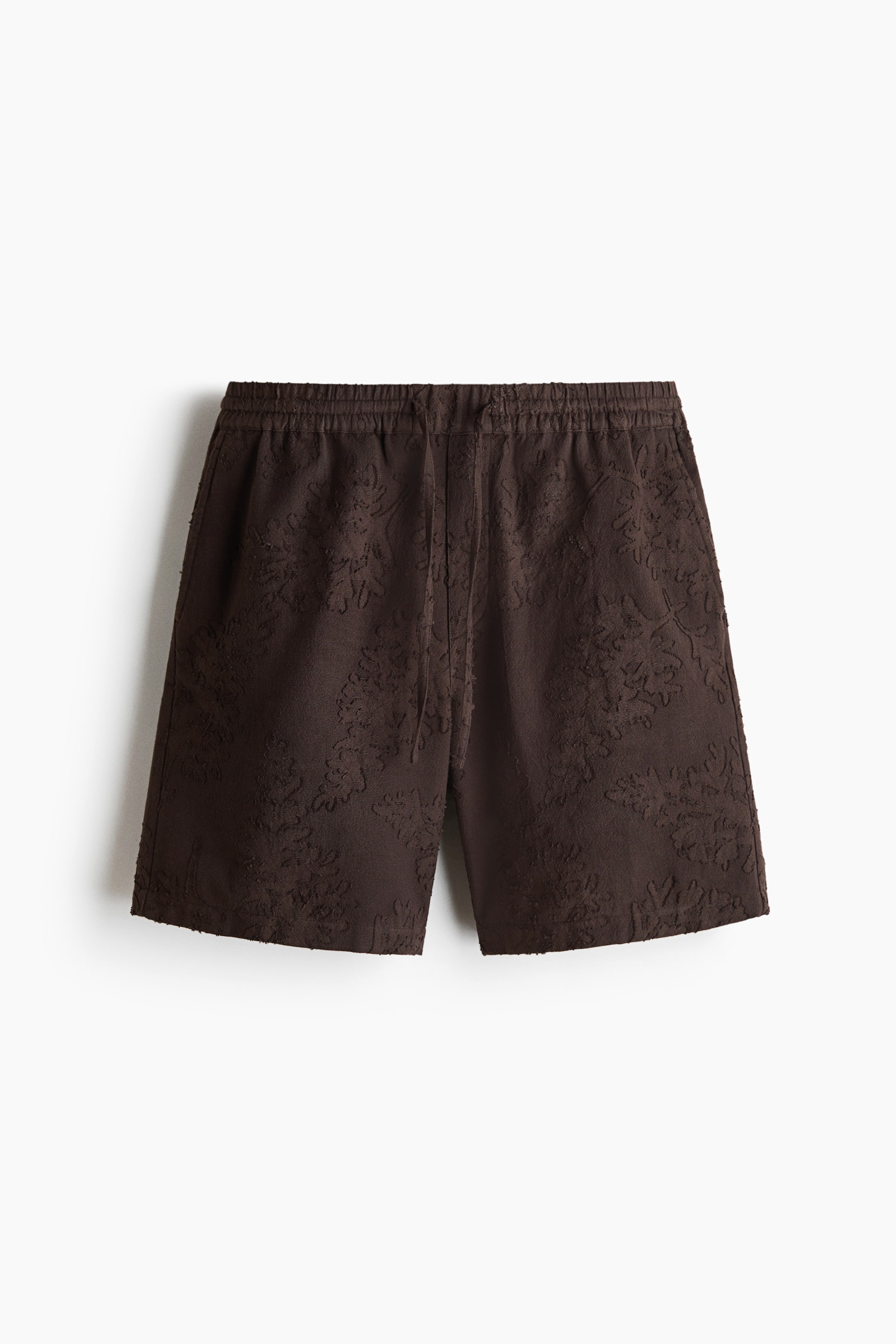 View larger image: Brown shorts are shown in a front view, showcasing an all-over dark brown embossed floral pattern. They feature an elasticated waistband with a visible drawstring, side pockets, and a raw hem.