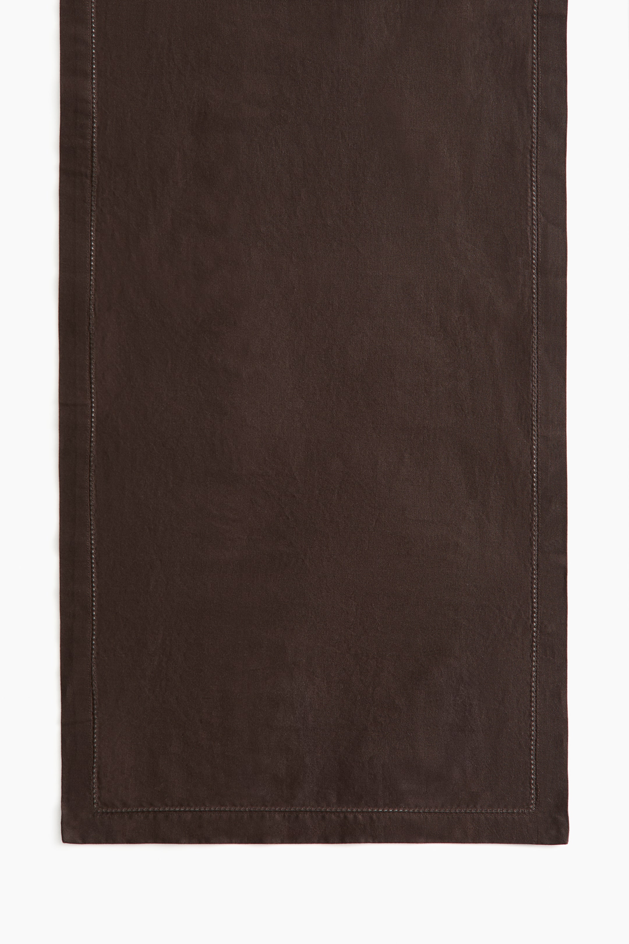 View larger image: A dark brown cotton-weave table runner is shown from the front, featuring textured fabric with delicate hemstitch detailing along the inner borders. The runner has a wide, folded hem finishing its outer edges.