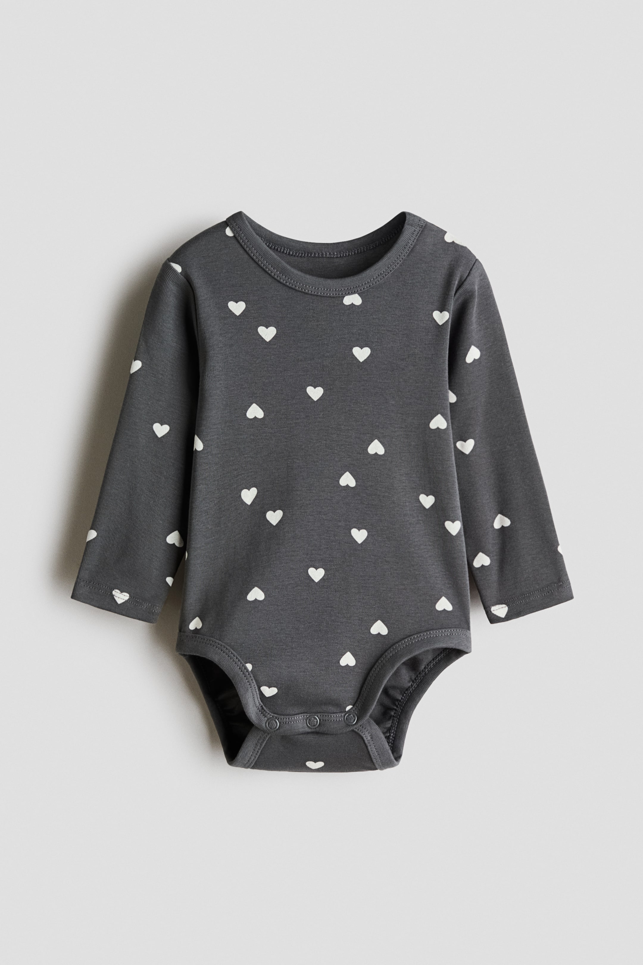 이미지 확대 보기: 앞면이 보이는 짙은 회색 저지 보디수트에는 흰색 하트 프린트가 있습니다. 라운드 트림 네크라인, 긴 소매, 한쪽 어깨의 프레스 스터드, 그리고 밑단의 숨겨진 프레스 스터드가 특징입니다.