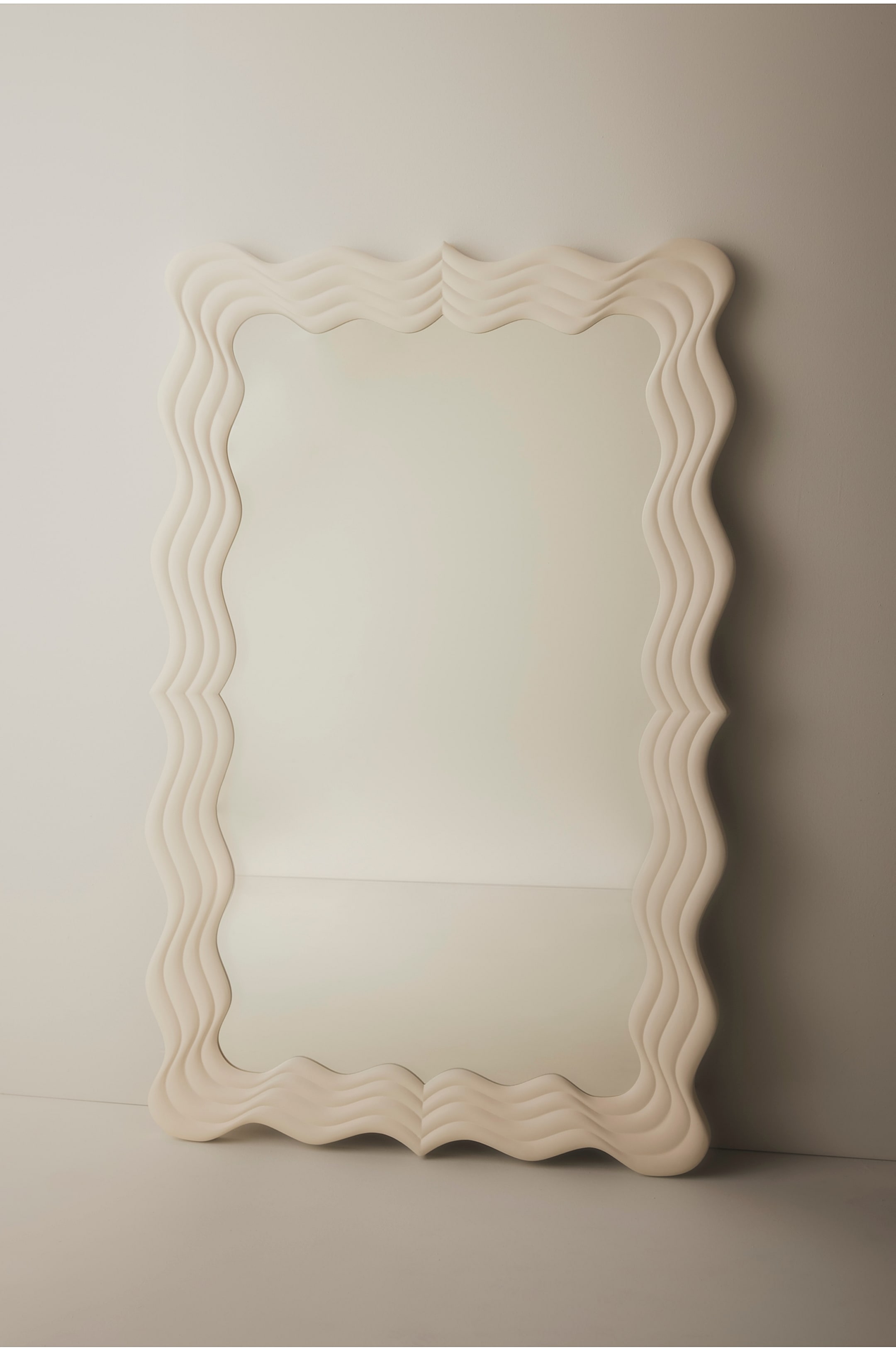 Agrandir l'image: Un grand miroir rectangulaire est incliné vers l’avant, mettant en valeur un large cadre couleur crème au design sculpté en vague ondulante. Le cadre présente des stries concentriques superposées qui s’incurvent autour de la vitre centrale réfléchissante.