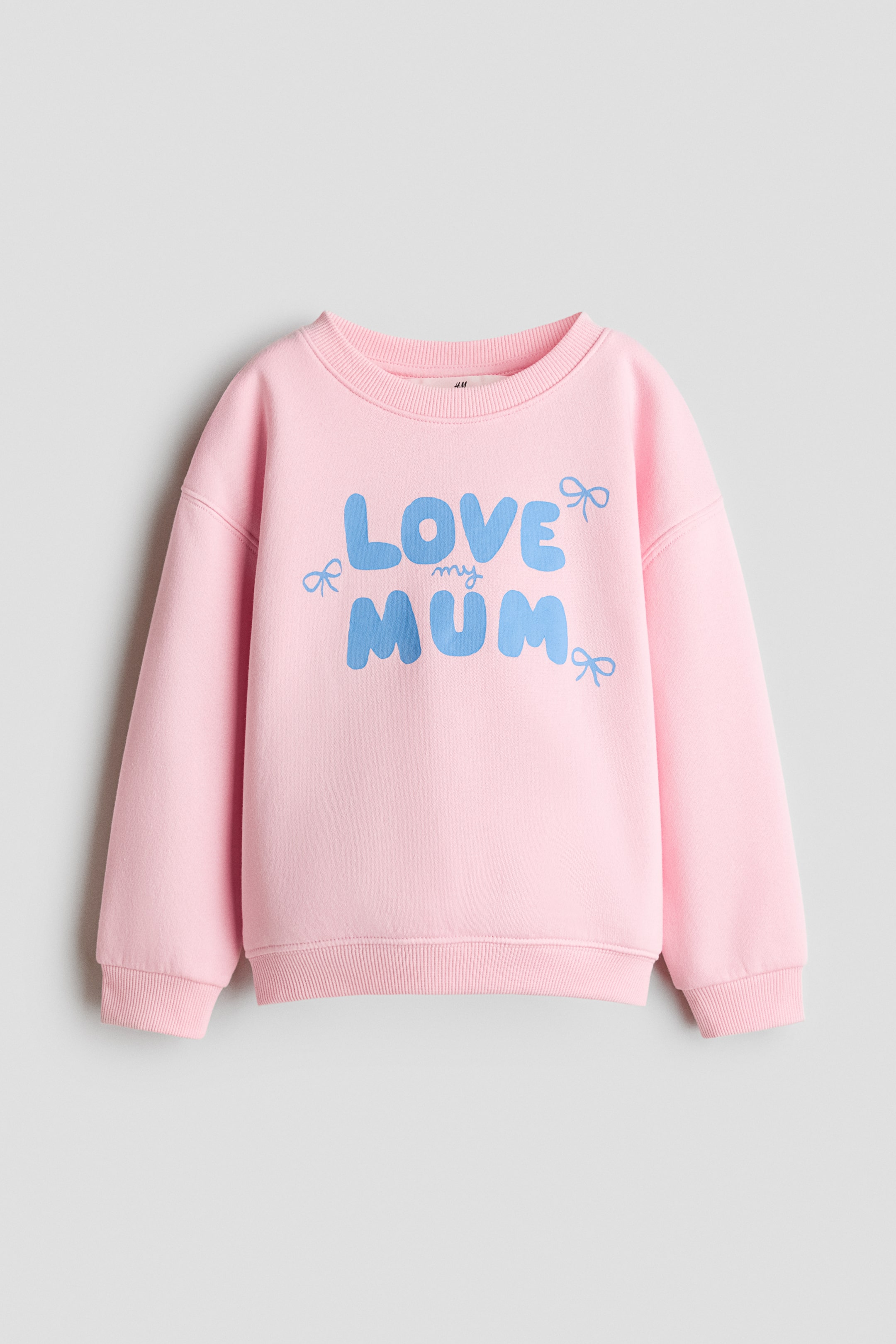View larger image: A pink long-sleeved sweatshirt is shown from the front, featuring a ribbed crew neck, dropped shoulders, ribbed cuffs, and a ribbed hem. The chest displays blue graphic text reading "LOVE my MUM" in bubble lettering, accented by three small blue bow illustrations.