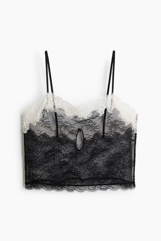 Een zwarte kanten camisole wordt recht van voren weergegeven, met dunne zwarte spaghettibandjes en een witte geschulpte kanten rand langs de bovenrand. Een sleutelgatuitsnijding is zichtbaar aan de voorkant in het midden, en de zoom onderaan is afgewerkt met zwarte geschulpte kant. - 1