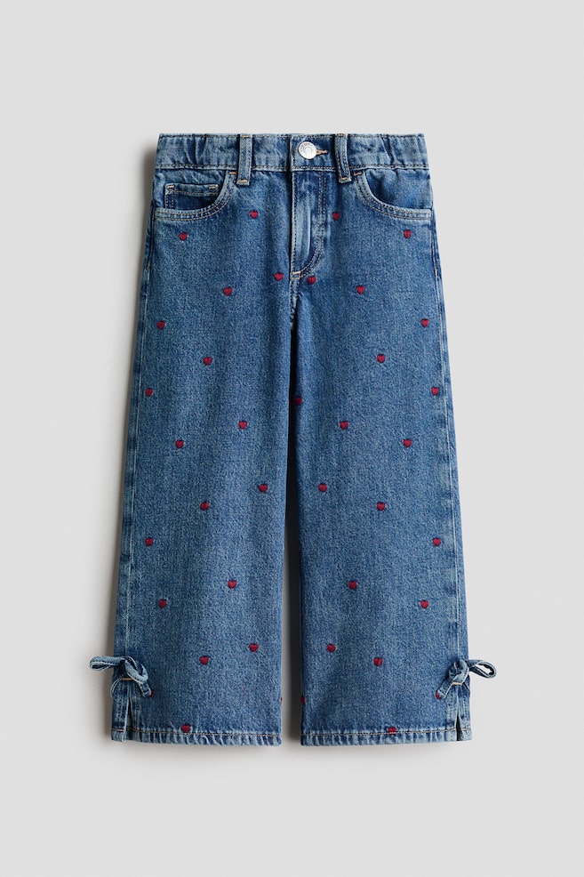A pair of blue cotton denim jeans is displayed from the front, featuring an all-over pattern of small, embroidered red hearts. The jeans are designed with a wide-leg fit, a five-pocket layout, an elasticated waistband, and small side slits at the bottom hems detailed with knotted denim ties. - 1