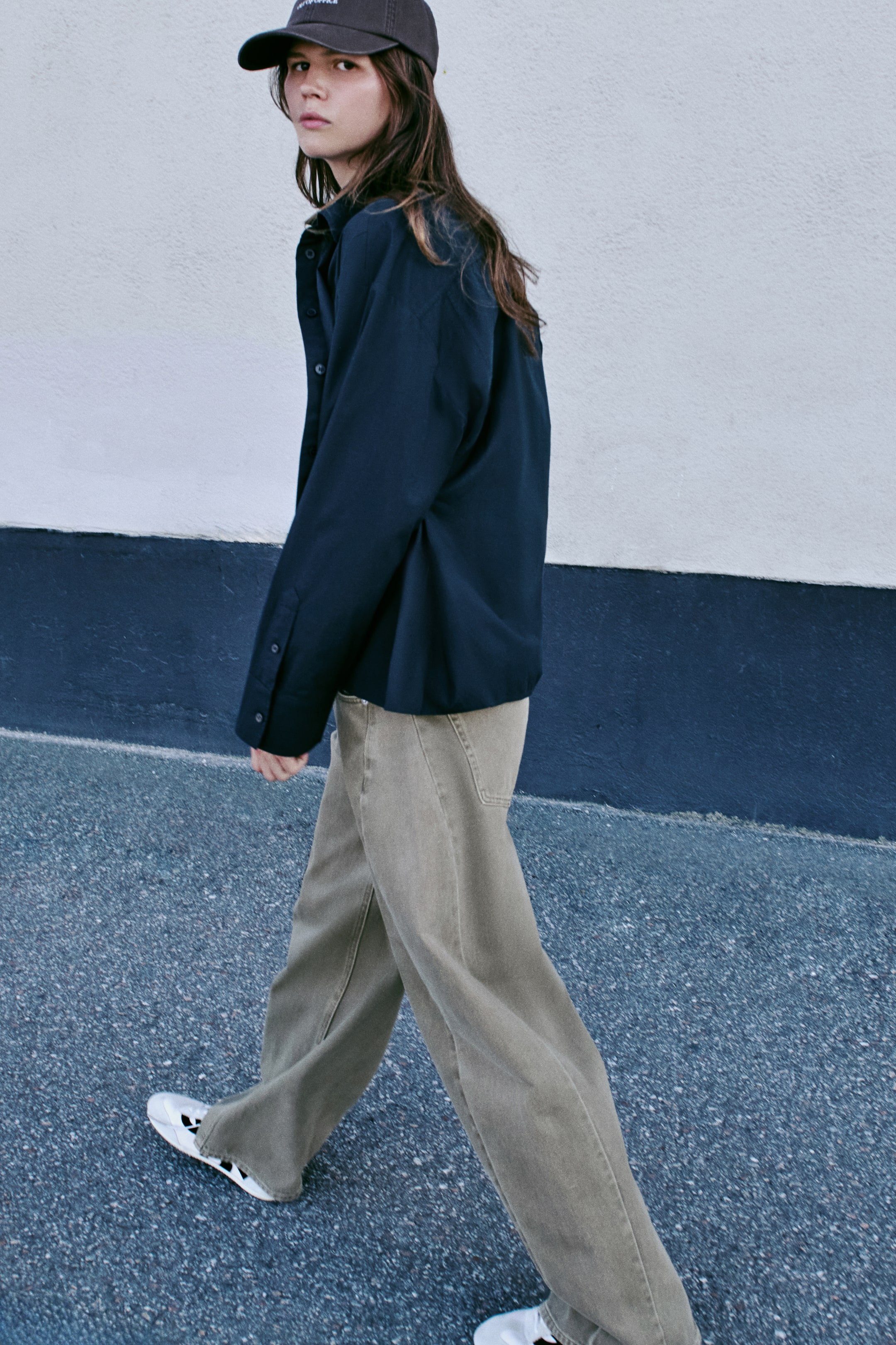 Visualizza l'immagine ingrandita: Una camicia in popeline di cotone blu navy scuro è mostrata di profilo laterale rivolto a sinistra, con colletto classico, spalle scese e maniche lunghe con polsini con bottoni. La camicia è indossata abbottonata, con una silhouette morbida che cade su pantaloni a gamba larga.