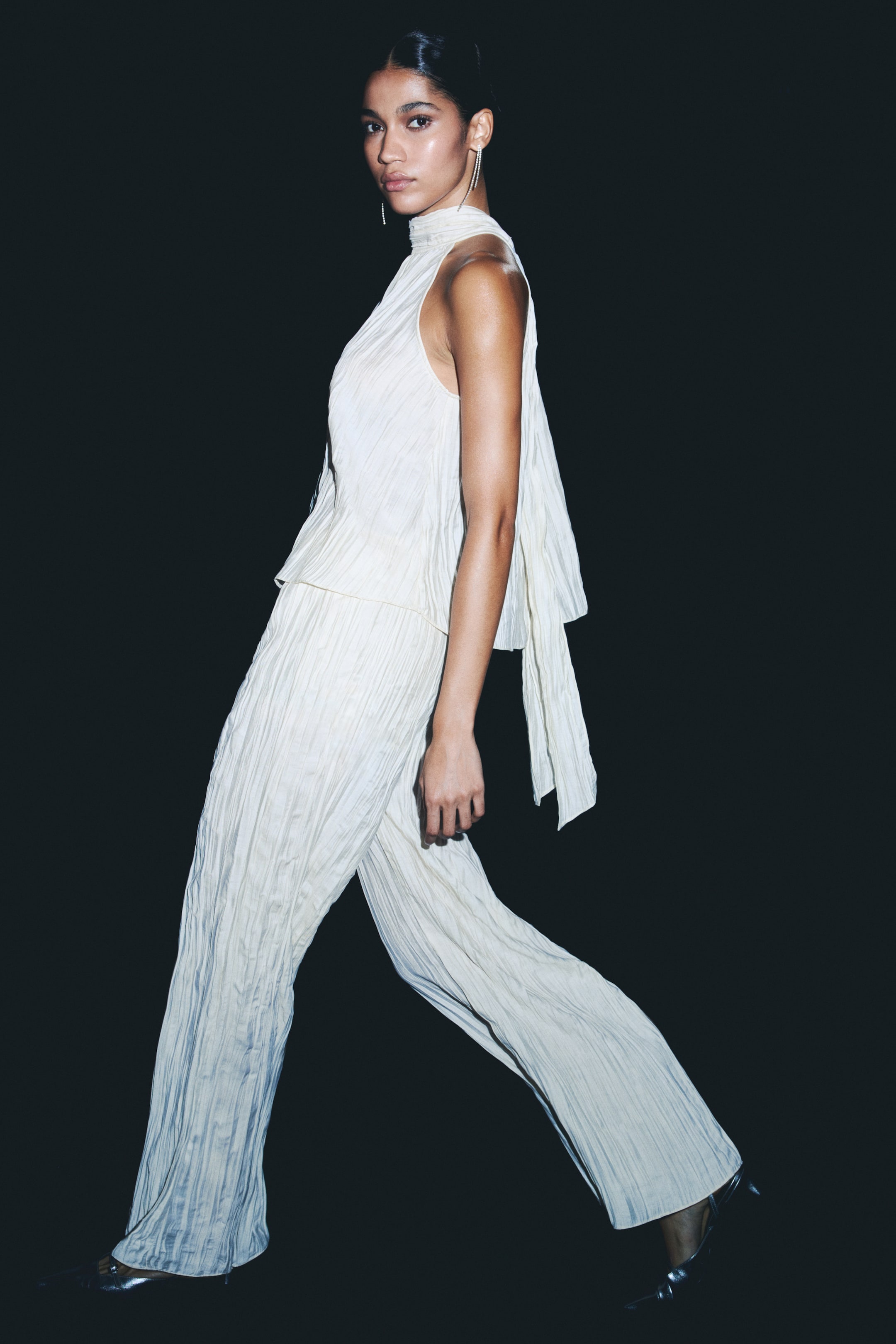 View larger image: A cream pleated two-piece set is facing left, with a sleeveless top featuring a high neck and a flowing, draped panel at the back. Wide-leg pants are completing the ensemble, and silver pointed-toe heels are being worn beneath their full-length hem.