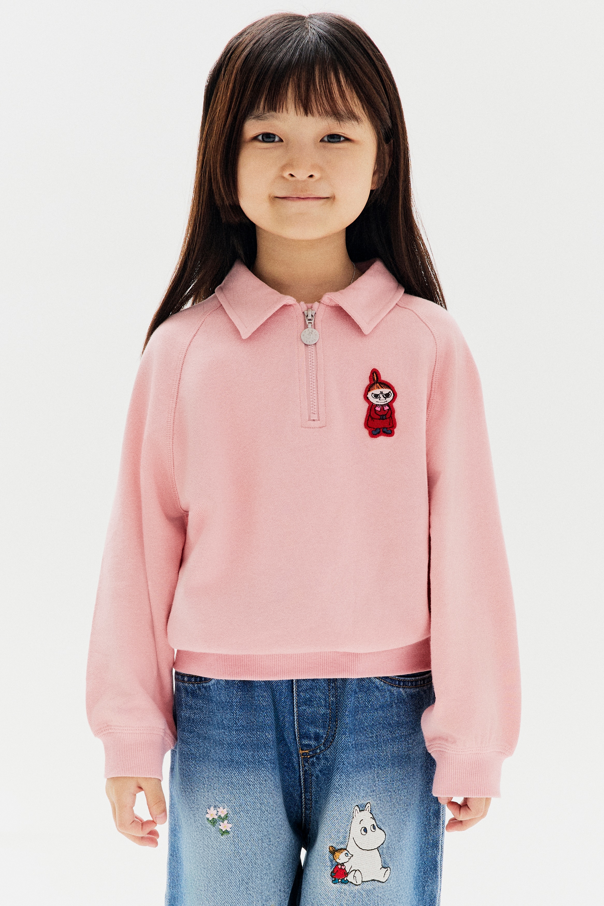 Má»t chiáº¿c Ã¡o sweatshirt mÃ u há»ng nháº¡t ÄÆ°á»£c chá»¥p tá»« phÃ­a trÆ°á»c, ná»i báº­t vá»i cá» Ã¡o polo vÃ  khÃ³a kÃ©o ná»­a thÃ¢n mÃ u báº¡c. Thiáº¿t káº¿ cÃ³ tay dÃ i raglan, gáº¥u Ã¡o vÃ  bo tay Ã¡o dá»t rib, cÃ¹ng má»t miáº¿ng patch thÃªu hÃ¬nh nhÃ¢n váº­t hoáº¡t hÃ¬nh nhá» á» ngá»±c trÃ¡i.