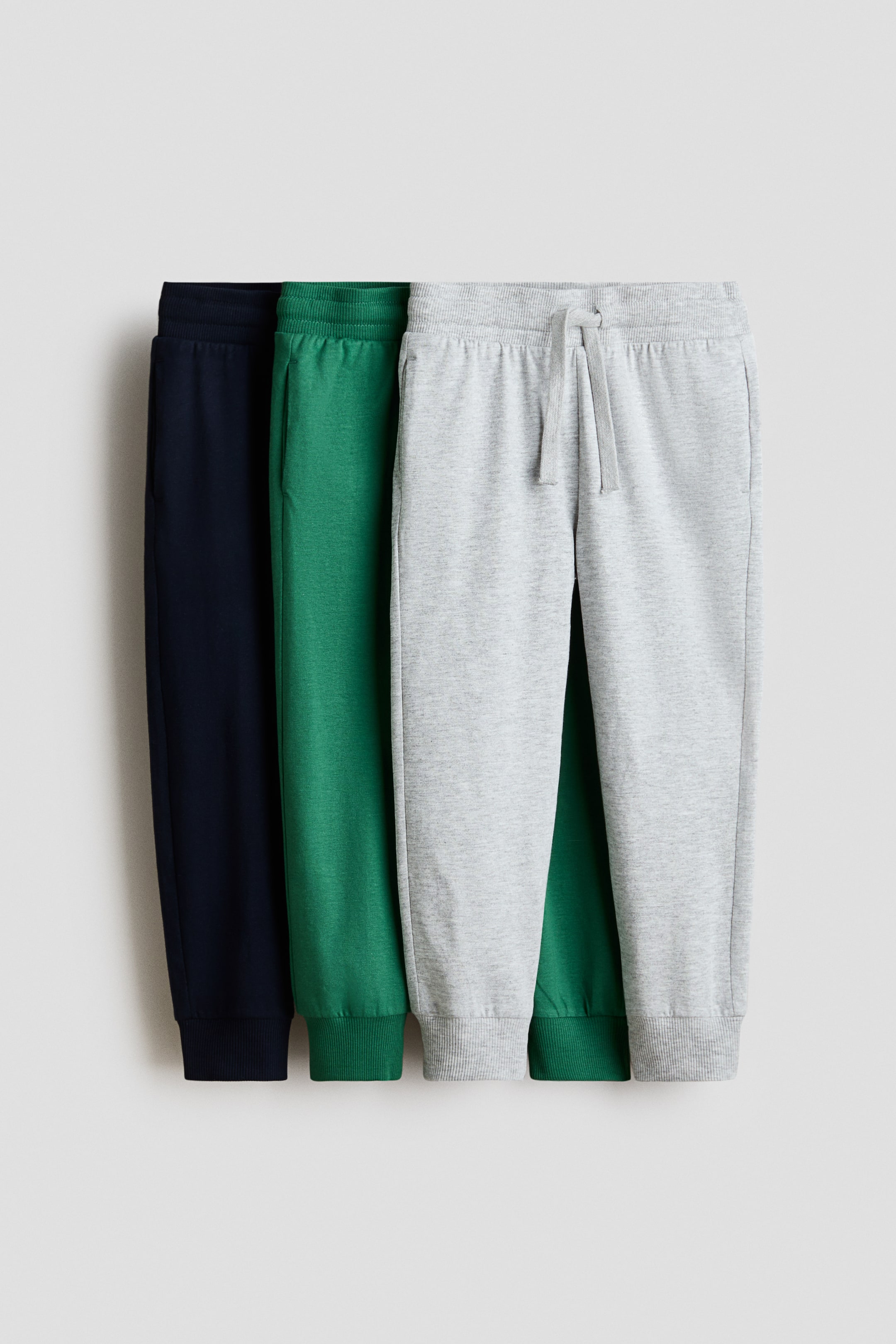 Ver imagem maior: Três pares de joggers em jersey de algodão macio — azul-escuro, verde e cinzento-claro — apresentados de frente, cada um com cintura elástica, bolsos nas costuras laterais e bainhas caneladas. O par cinzento-claro apresenta ainda um cordão na cintura.