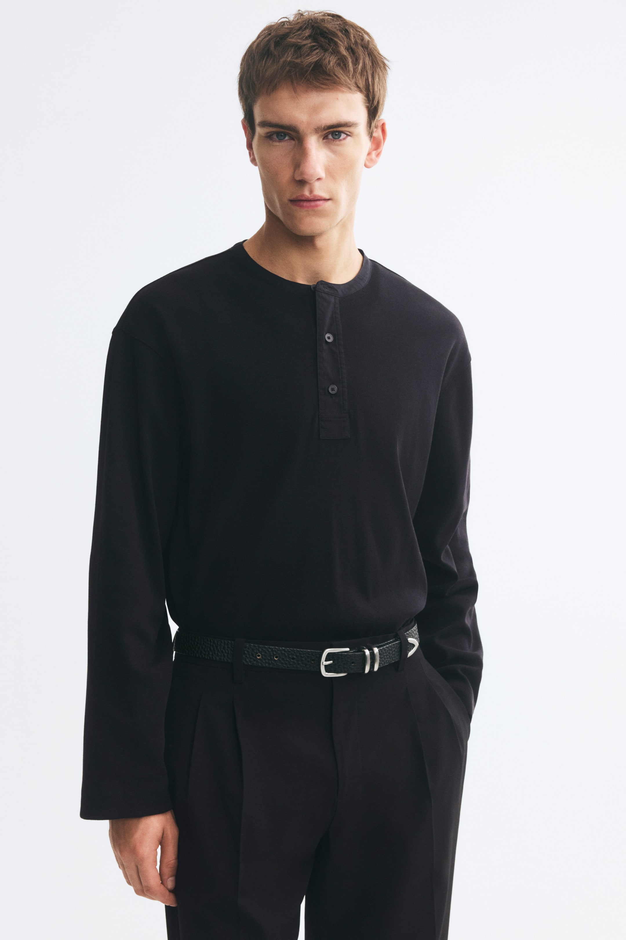 Grotere afbeelding bekijken: Een zwarte Henley-shirt met lange mouwen in ribgebreide stof wordt gedragen door het model, ingestopt in een zwarte broek die is vastgezet met een riem met textuur. Het shirt heeft een relaxed fit met verlaagde schouders, een ronde halslijn en een gedeeltelijk geknoopte knoopsluiting aan de voorkant.