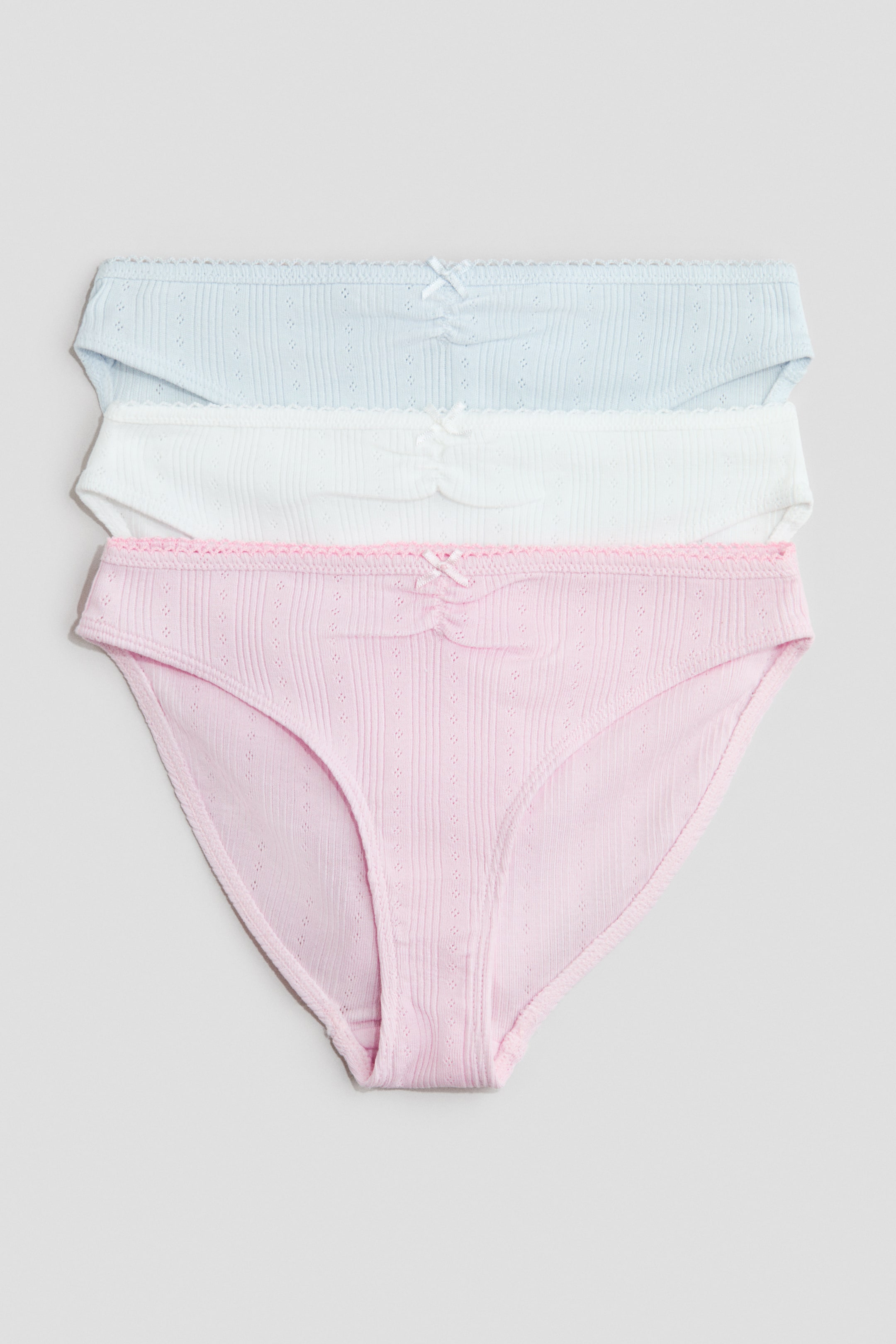 Ingrandisci l'immagine: Tre slip in jersey di cotone sono esposti in vista frontale mentre sono impilati. Gli slip, nei colori azzurro chiaro, bianco e rosa chiaro, presentano arricciature e un piccolo fiocco decorativo al centro della cintura anteriore, e sono rifiniti con un delicato bordino lungo la parte superiore.