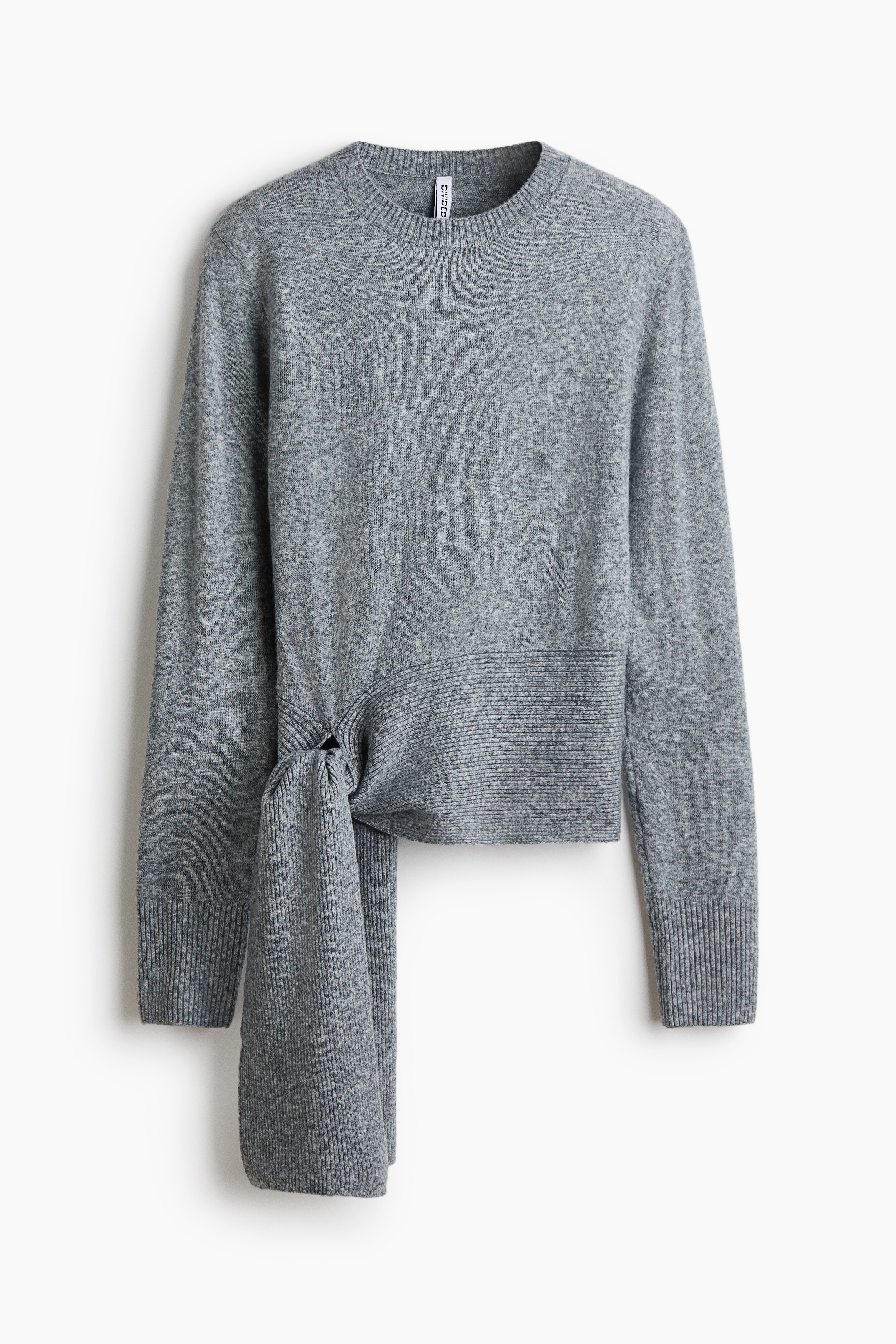 La modelo lleva un jersey de punto fino gris, con cuello redondo acanalado, mangas largas con puÃ±os acanalados y un bajo con lazadas anchas anudadas en el lateral delantero. El jersey se combina con unos vaqueros azul claro y un cinturÃ³n negro, dejando ver parte de la cintura.