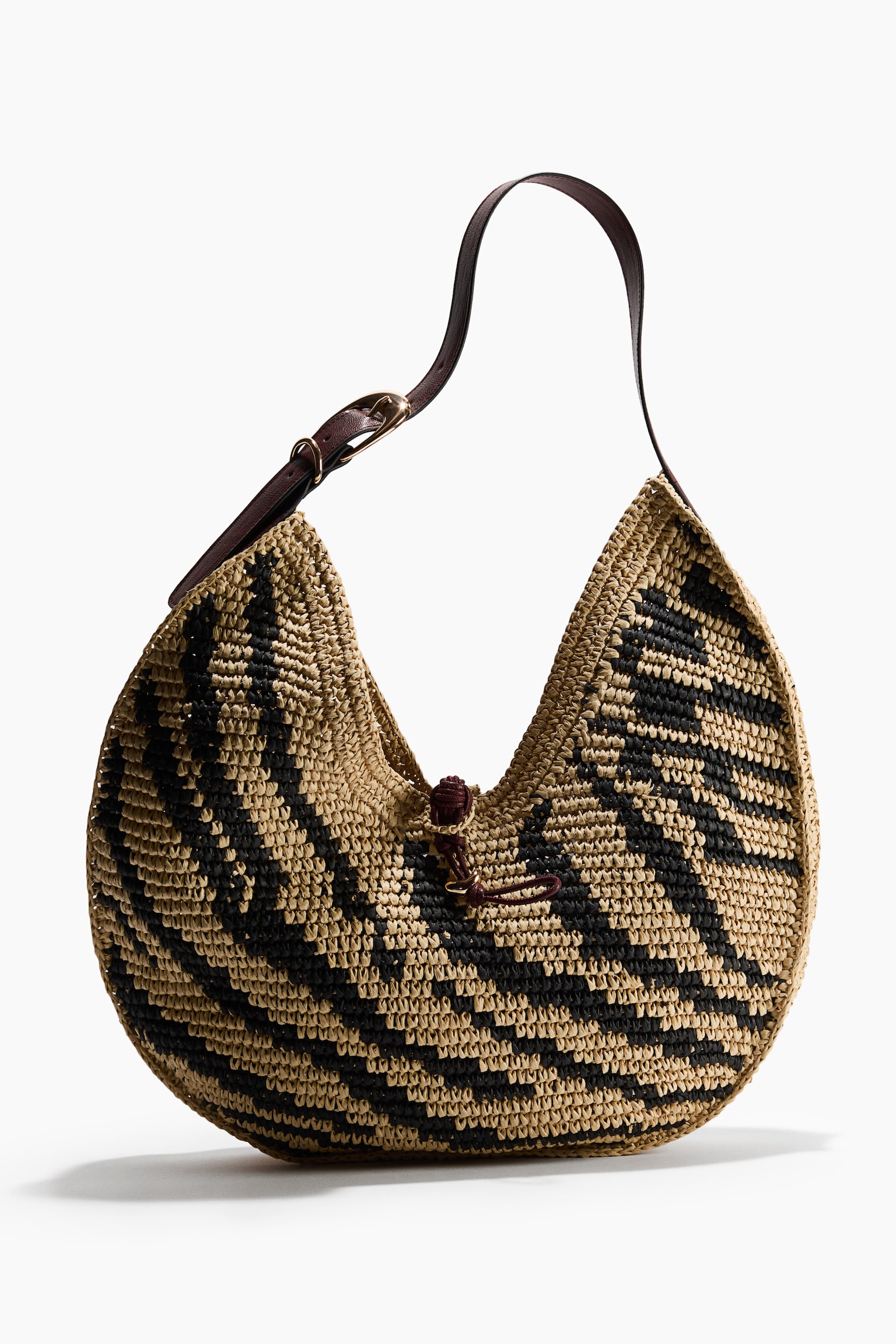 A woven raffia hobo bag showcases a beige and black zigzag pattern. A dark brown leather strap extends from the top, fastened by a brass-toned buckle, while a small dark brown cord cinches the central opening. - 1