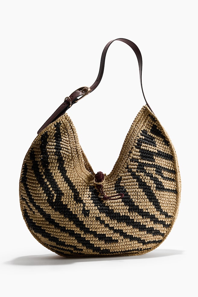 A woven raffia hobo bag is featuring a beige and black zigzag pattern. A dark brown leather strap is extending from the top, secured by a brass-toned buckle, and a small dark brown cord is cinching the central opening. - 1