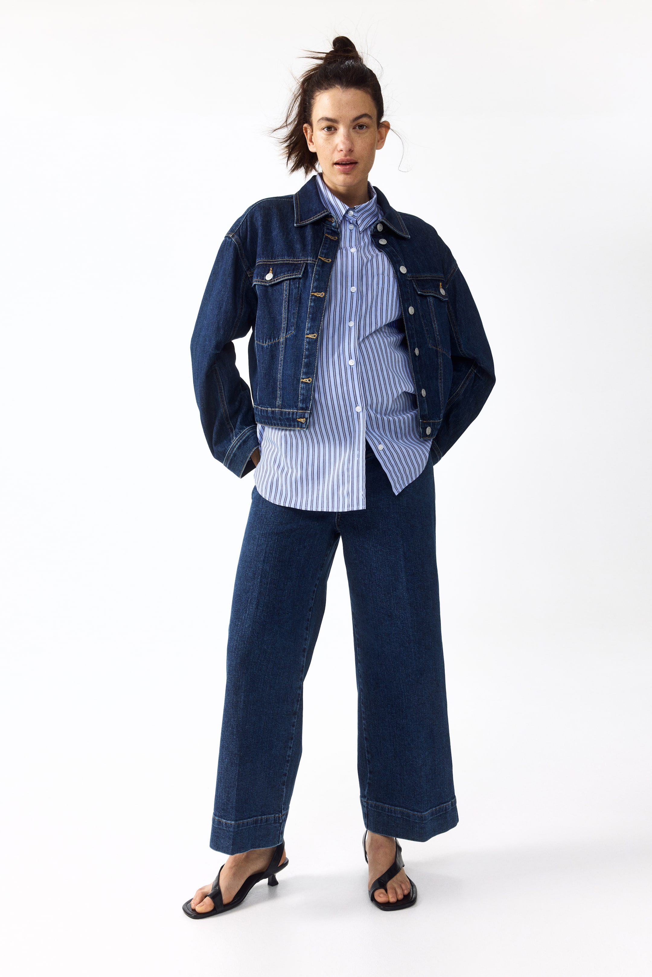 Zobraziť väčší obrázok: Tmavá džínsová bunda s uvoľneným strihom, ktorá má gombíkové náprsné vrecká a špicatý golier, je nosená na modro-bielej vertikálne pruhovanej košeli s gombíkmi. Tmavé džínsy so širokými nohavicami a ohrnutými lemami dopĺňajú celkový vzhľad, všetko prezentované z predného pohľadu.