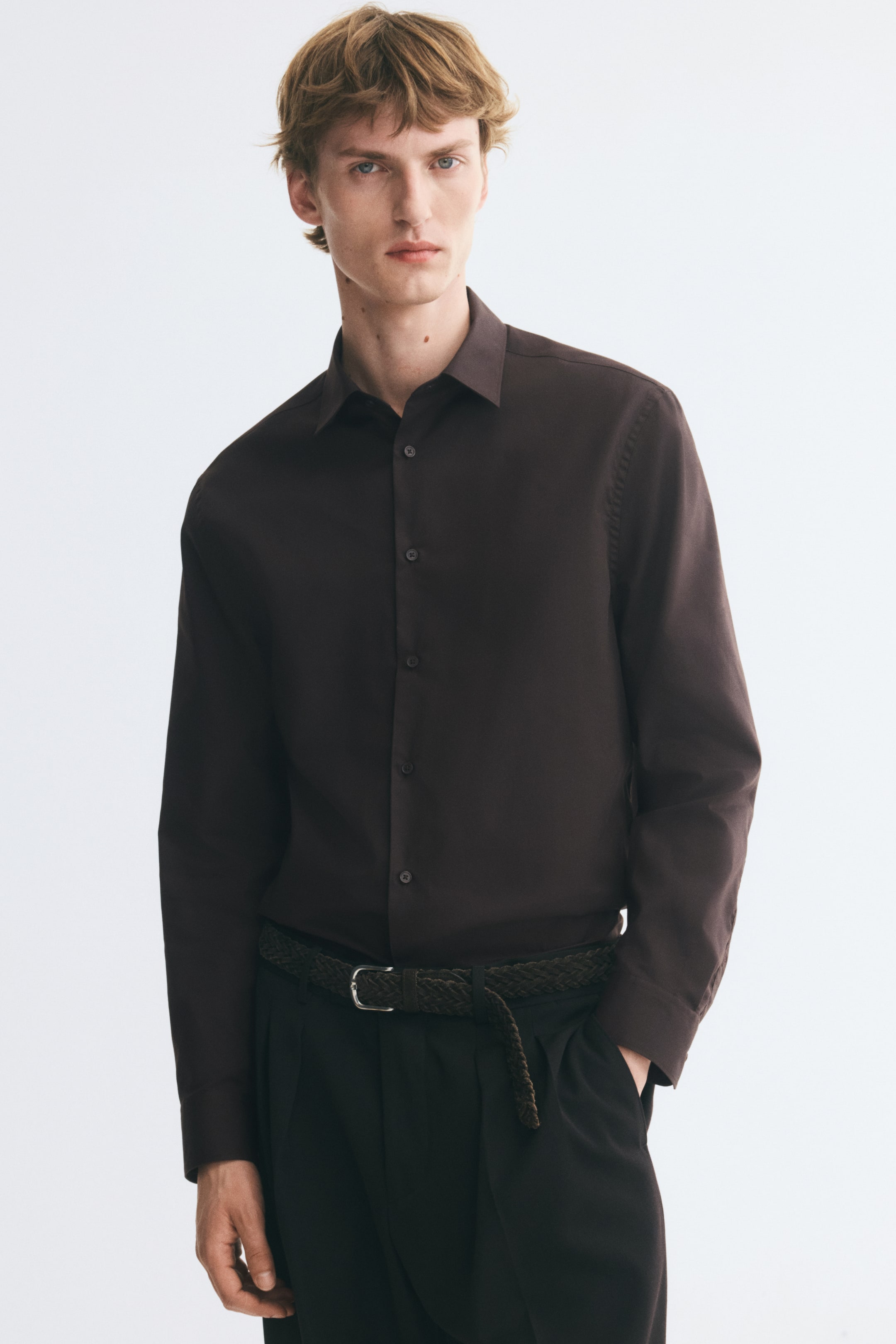 View larger image: The model is wearing a dark brown woven shirt with a turn-down collar and long sleeves, tucked into dark trousers with a braided belt. The shirt features a regular fit, a button-up front, and buttoned cuffs.