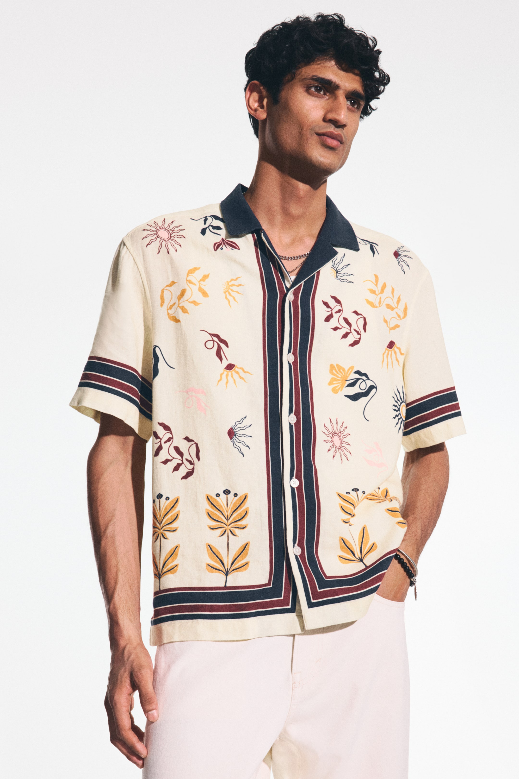 View larger image: The model is wearing a short-sleeved, cream-coloured printed shirt, showcasing a dark blue resort collar, a French front with a central striped panel in dark blue and maroon, and short sleeves finished with matching striped bands. The shirt is embellished with vibrant abstract botanical patterns. The model pairs it with light-coloured trousers, casually placing one hand in a pocket.