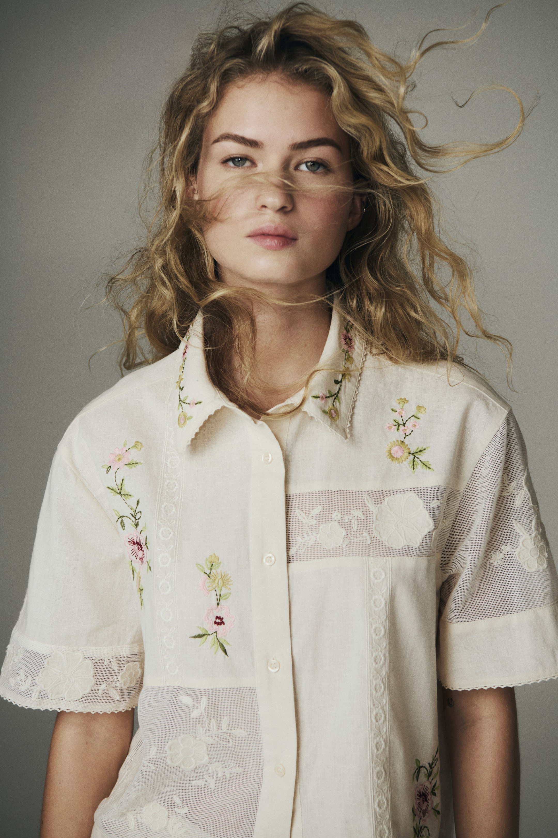 Grotere afbeelding bekijken: Een crèmewitte blouse met korte mouwen en een kraag, voorzien van bloemenborduursels, kanten inzetten en doorschijnende geborduurde panelen, wordt door het model gedragen.