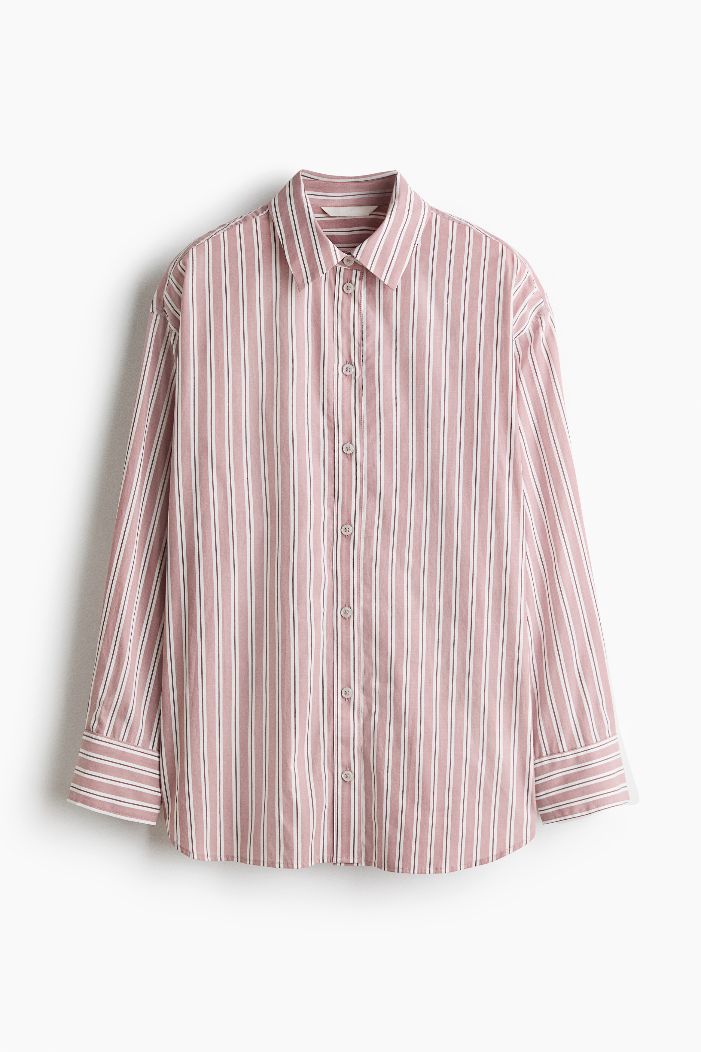 An oversized pink-and-white vertically striped cotton shirt is worn open by the model, layered over a white tank top and paired with wide-leg off-white trousers. The shirt features dropped shoulders, a collar, and long sleeves with folded cuffs.