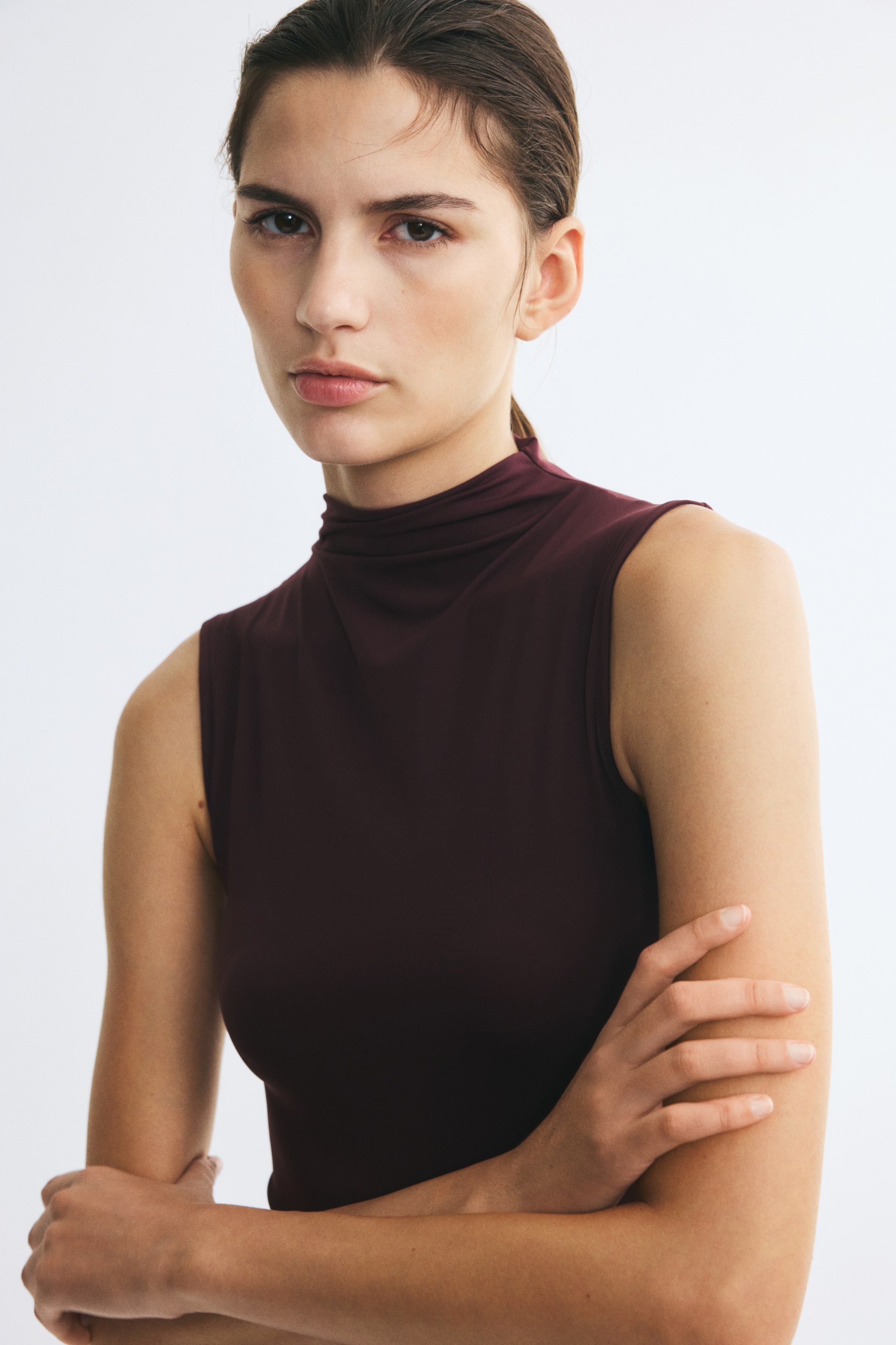 Voir une image plus grande: Un body sans manches, de couleur bourgogne foncée, doté d’un col roulé haut, est porté par le mannequin, qui pose les bras croisés.