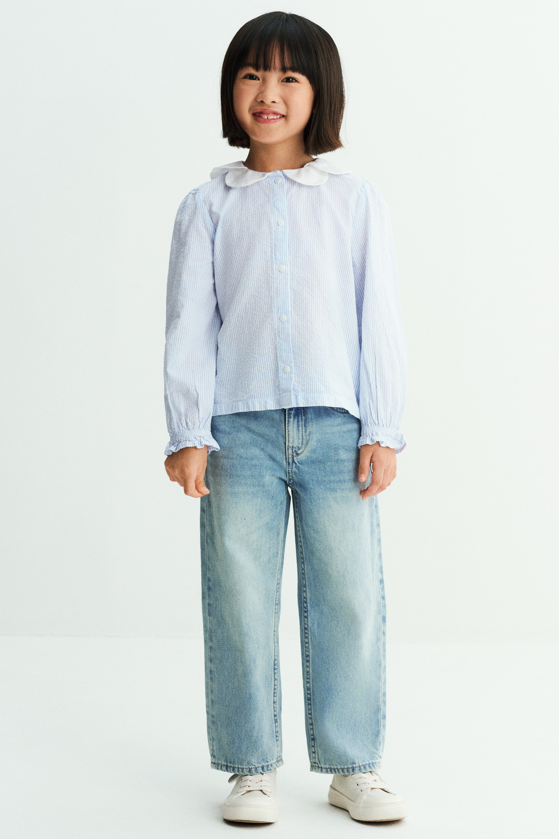 View larger image: The model is wearing a light blue and white striped seersucker blouse, featuring a white rounded two-piece collar, button-down front, and long puff sleeves with elasticated flounced cuffs. It is styled with wide-leg light-wash denim jeans.