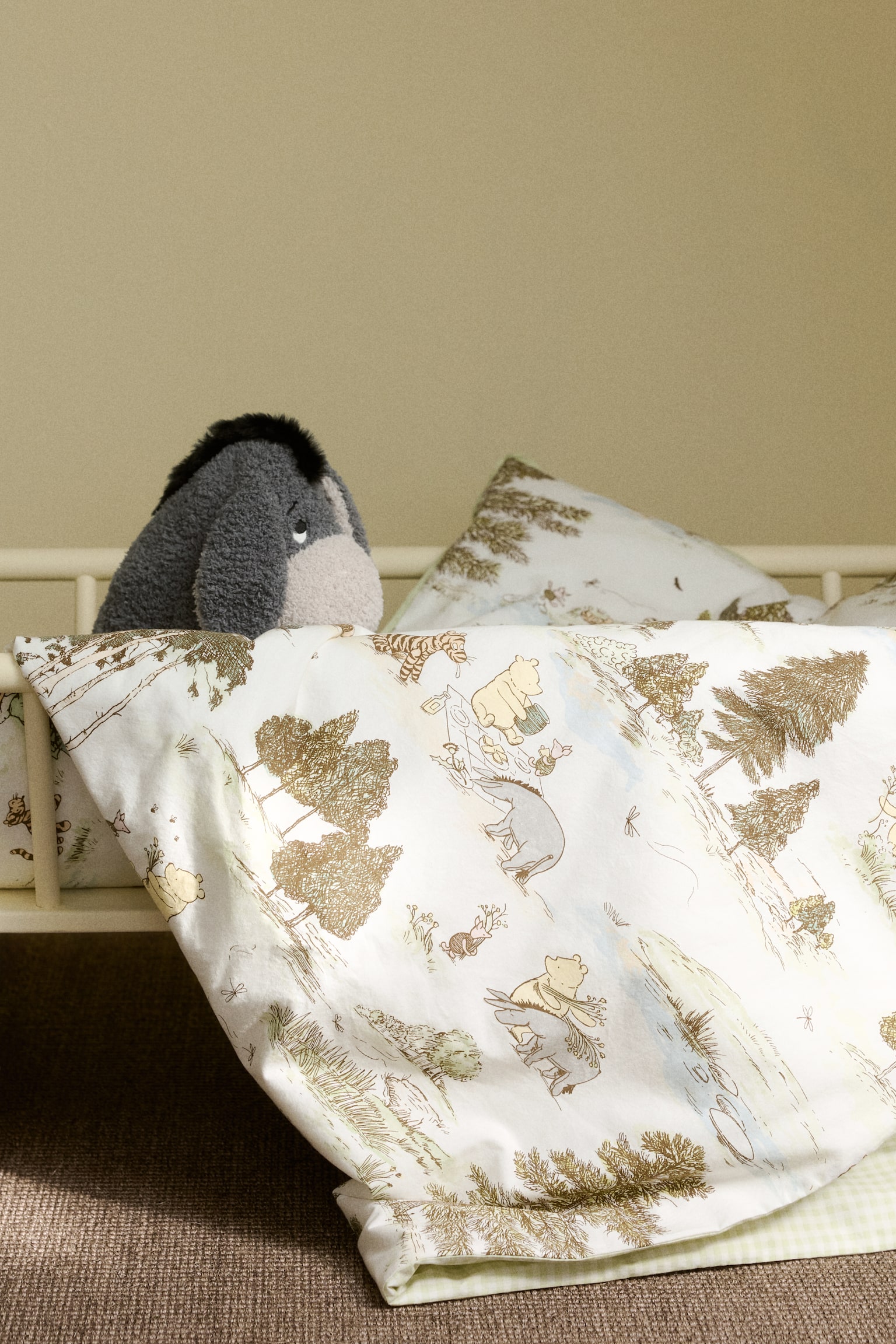 The bed linen features a white duvet cover and pillowcase adorned with an illustrated Winnie the Pooh pattern. The design showcases characters such as Pooh and Eeyore amidst brown trees and green foliage, accented with light blue tones. The duvet cover is elegantly draped over a bed frame, revealing a light green gingham pattern on its underside. - 1