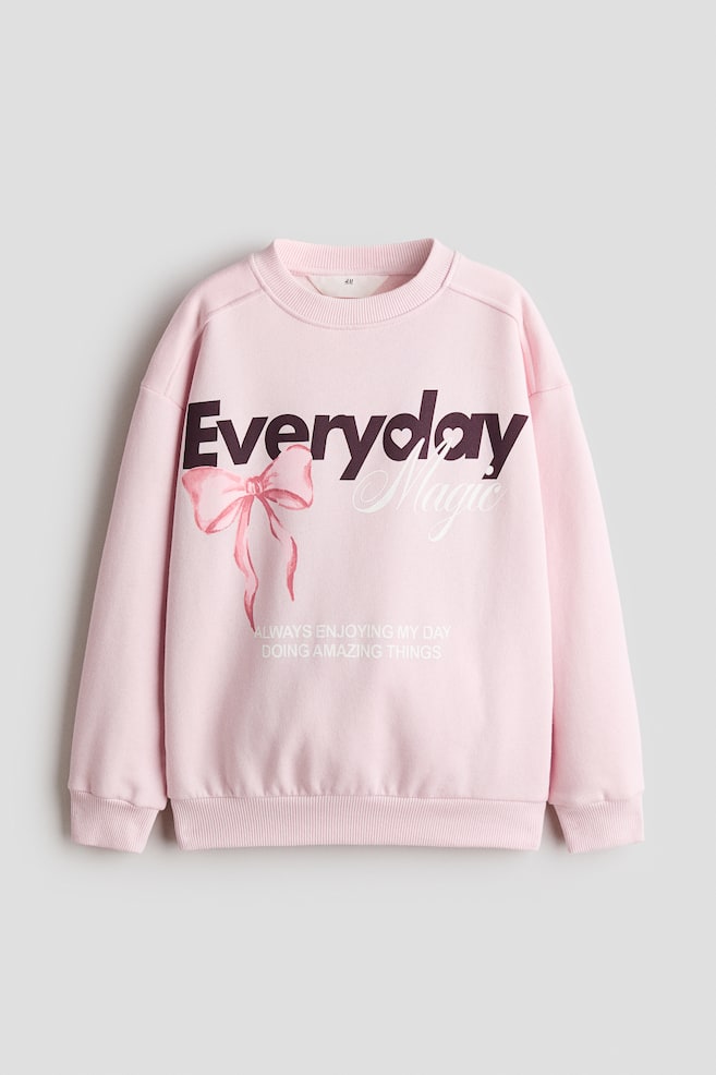 Ce sweat-shirt ample rose pâle, vu de face, présente un col rond bordé de côtes, des coutures dâépaules orientées vers lâavant et des épaules tombantes avec des manches longues. La poitrine arbore un motif imprimé avec les mots « Everyday Magic » en lettres violet foncé et blanches, accompagné dâune illustration de nÅud rose et dâun petit texte blanc en dessous. - 1