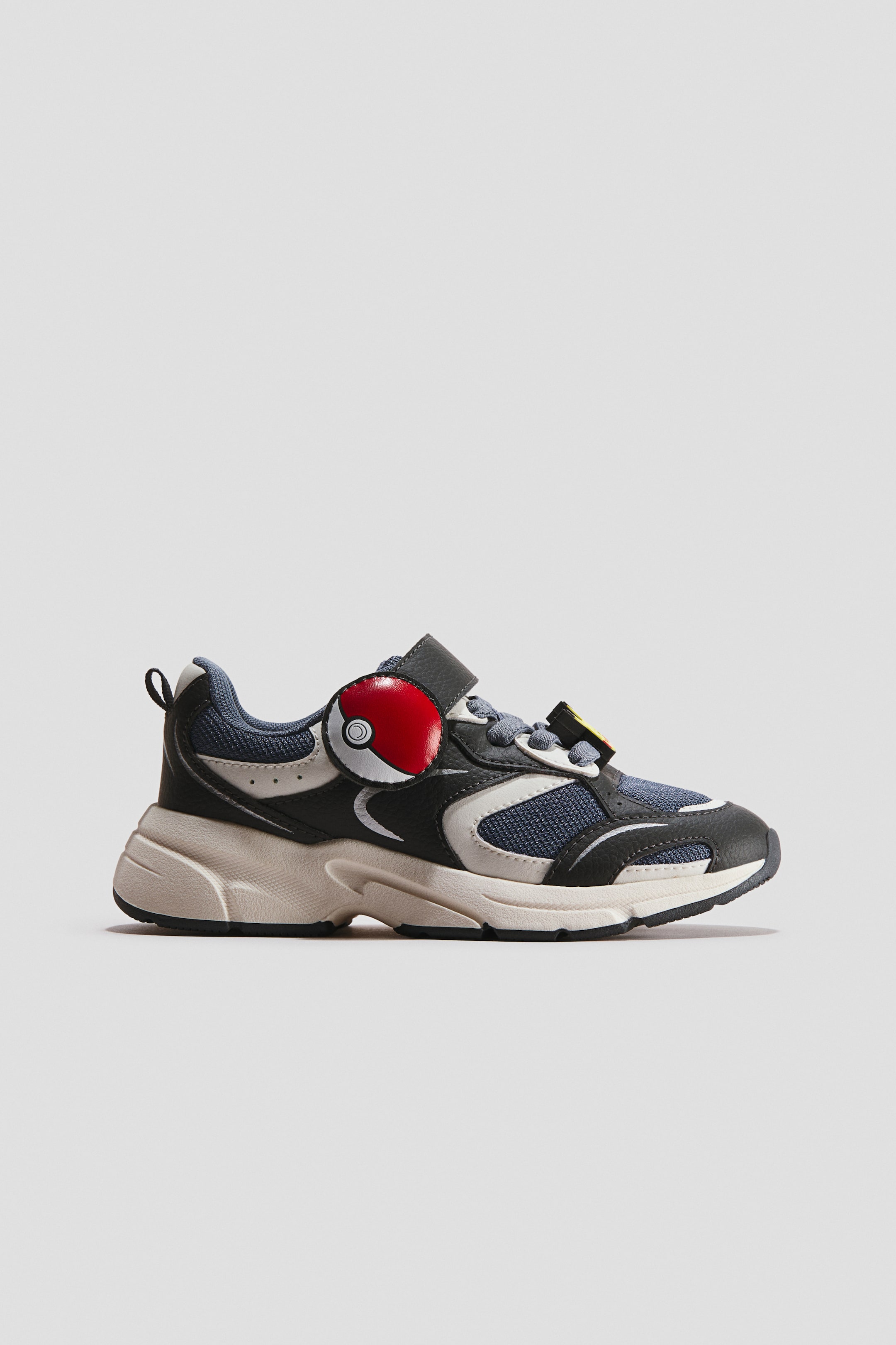 View larger image: A motif-detail trainer is shown in side profile, highlighting a paneled upper with black, grey, and blue mesh inserts. The shoe features a chunky off-white sole, grey laces, and a hook-and-loop strap adorned with a round red-and-white circular motif.