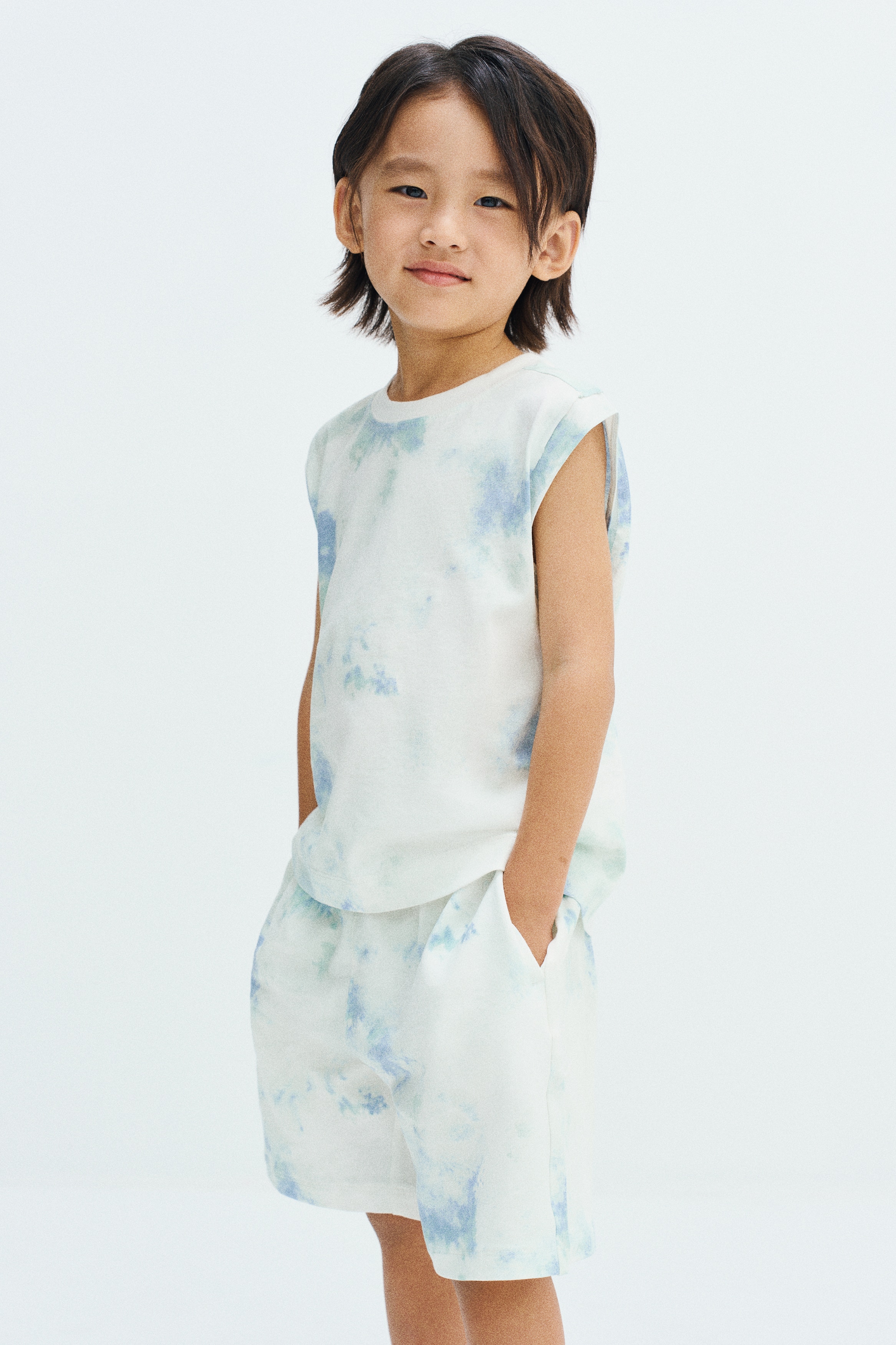 A sleeveless t-shirt, front-facing, is displayed with a white base decorated with a soft blue and light green tie-dye pattern. The garment features a ribbed crewneck, wide armholes, and a straight hem.