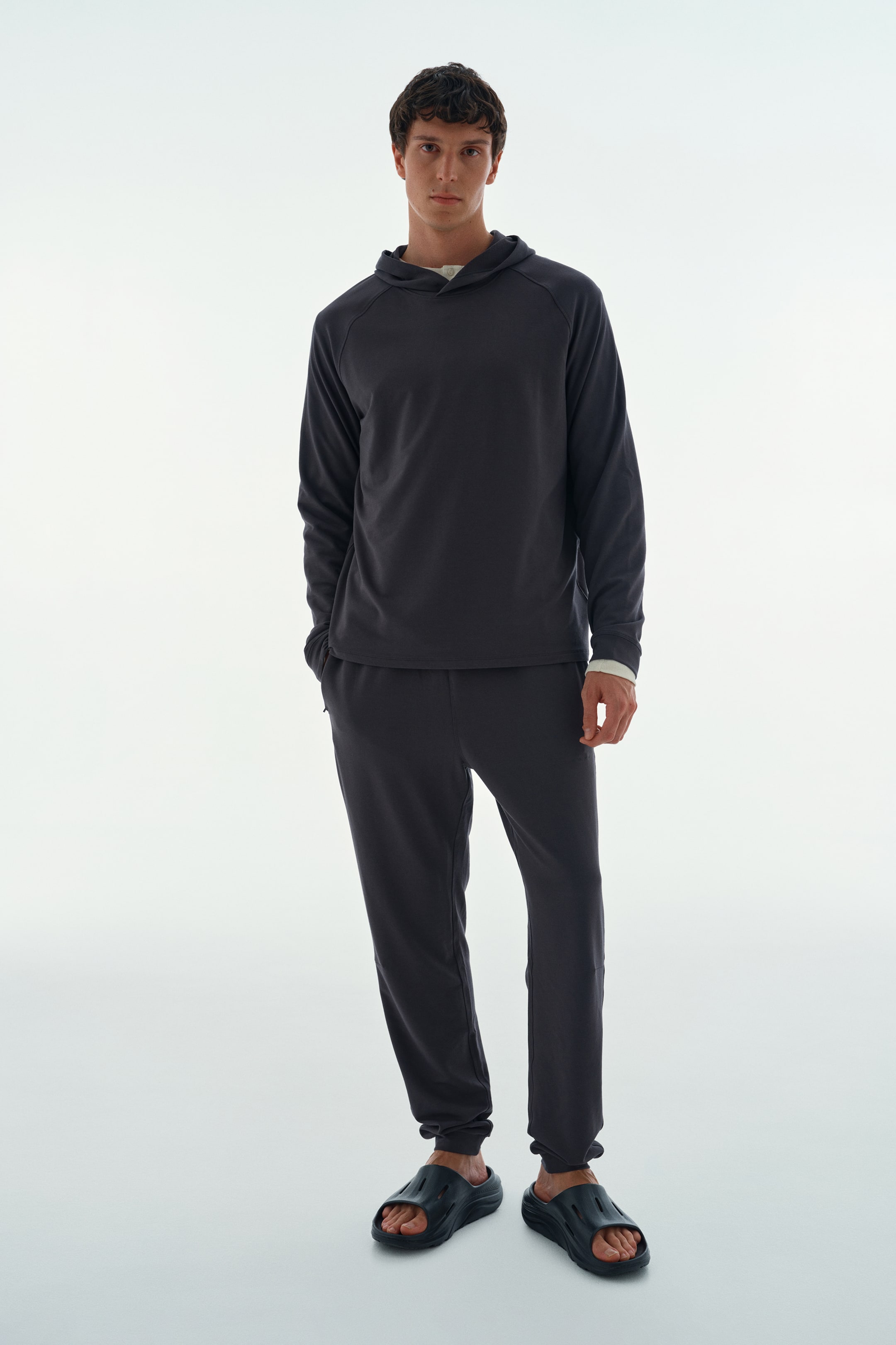 Näytä suurempi kuva: Malli käyttää tummanharmaata loungewear-settiä, johon kuuluu pitkähihaista hupparia ja siihen sopivat jogger-housut. Hupparin alla on vaalea paita, joka näkyy kauluksesta ja hihansuista, ja malli seisoo toinen käsi housuntaskussa.