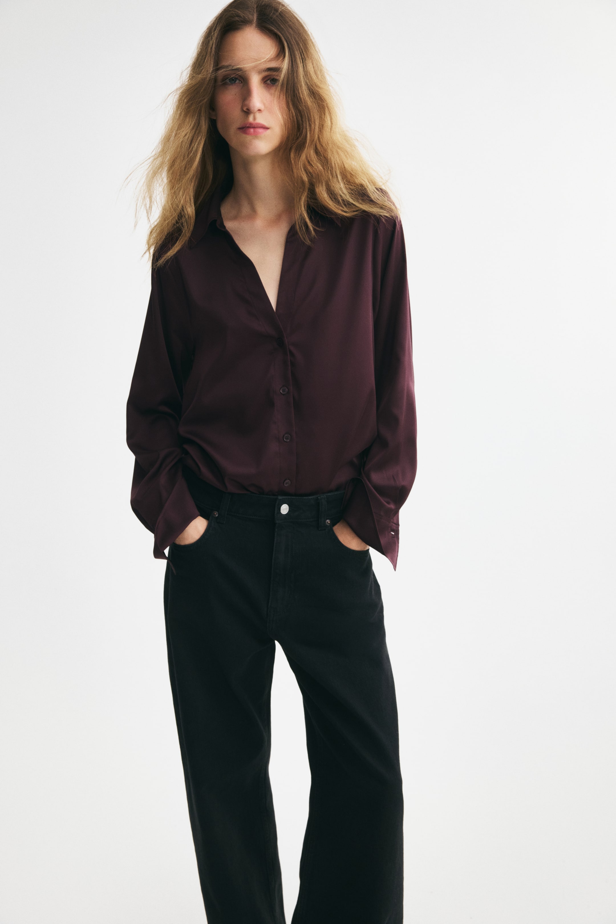 Grösseres Bild ansehen: Das Model ist von vorne zu sehen und trägt eine tief burgunderfarbene Bluse mit V-Ausschnitt aus gewebtem Stoff mit dezentem Glanz, kombiniert mit in die schwarzen Jeans gesteckter Bluse. Die Bluse verfügt über einen klassischen Kragen, eine durchgehende Knopfleiste und lange, lockere Ärmel mit breiten Manschetten mit Knöpfen.