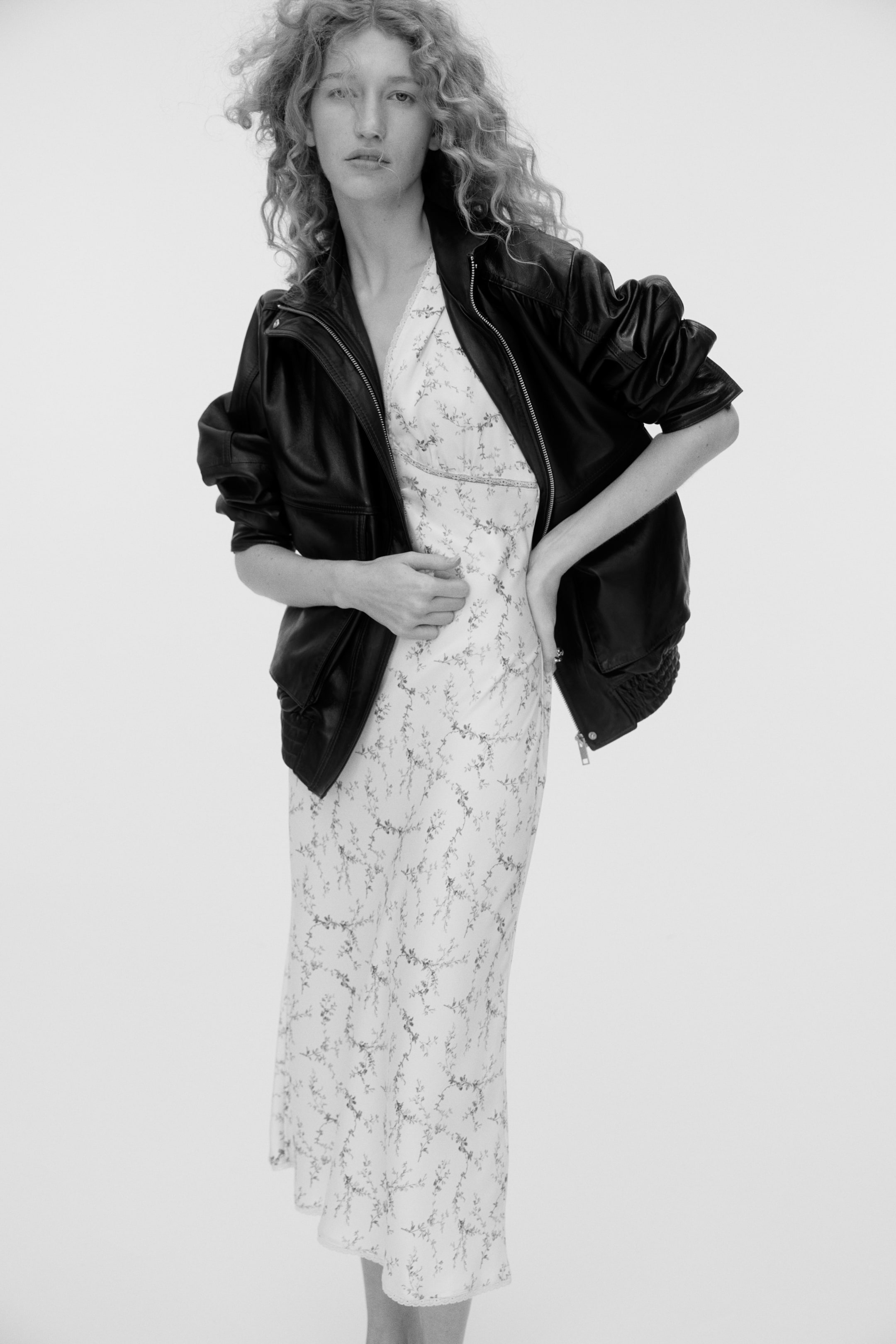 View larger image: A black leather bomber jacket is worn open over a white midi slip dress, shown from a front-facing angle. The jacket features a zippered front and gathered sleeves, while the slip dress displays a delicate floral pattern and a V-neckline.