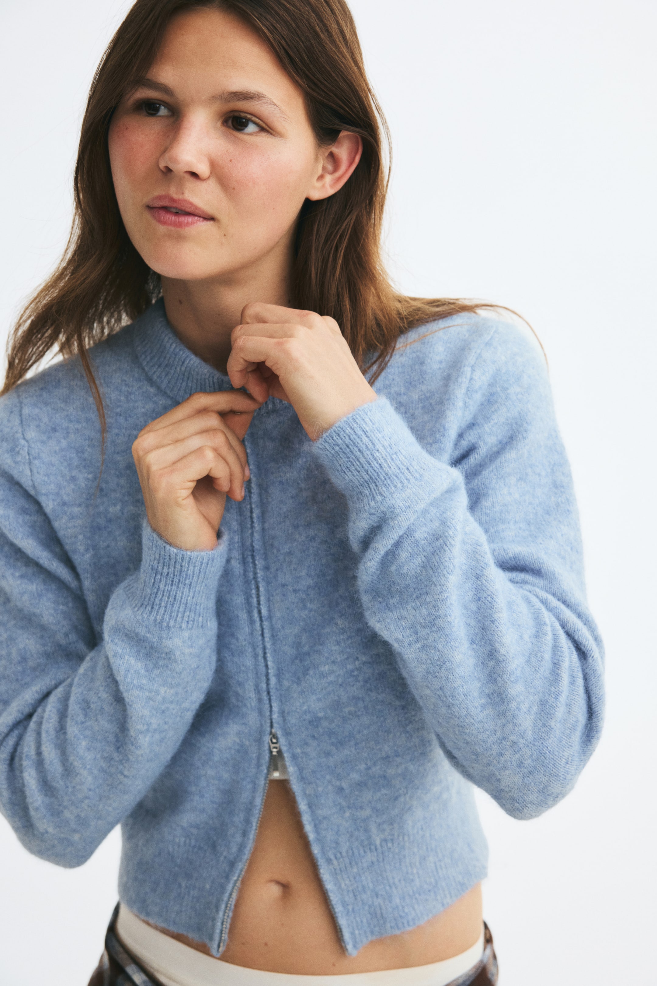 Vezi imaginea mai mare: Un cardigan din tricot moale, de culoare albastru deschis, cu decolteu rotund cu model rib, fermoar frontal bidirecțional și manșete rib este purtat de model. Modelul ajustează fermoarul de la guler cu ambele mâini, lăsând partea de jos a cardiganului parțial deschisă.