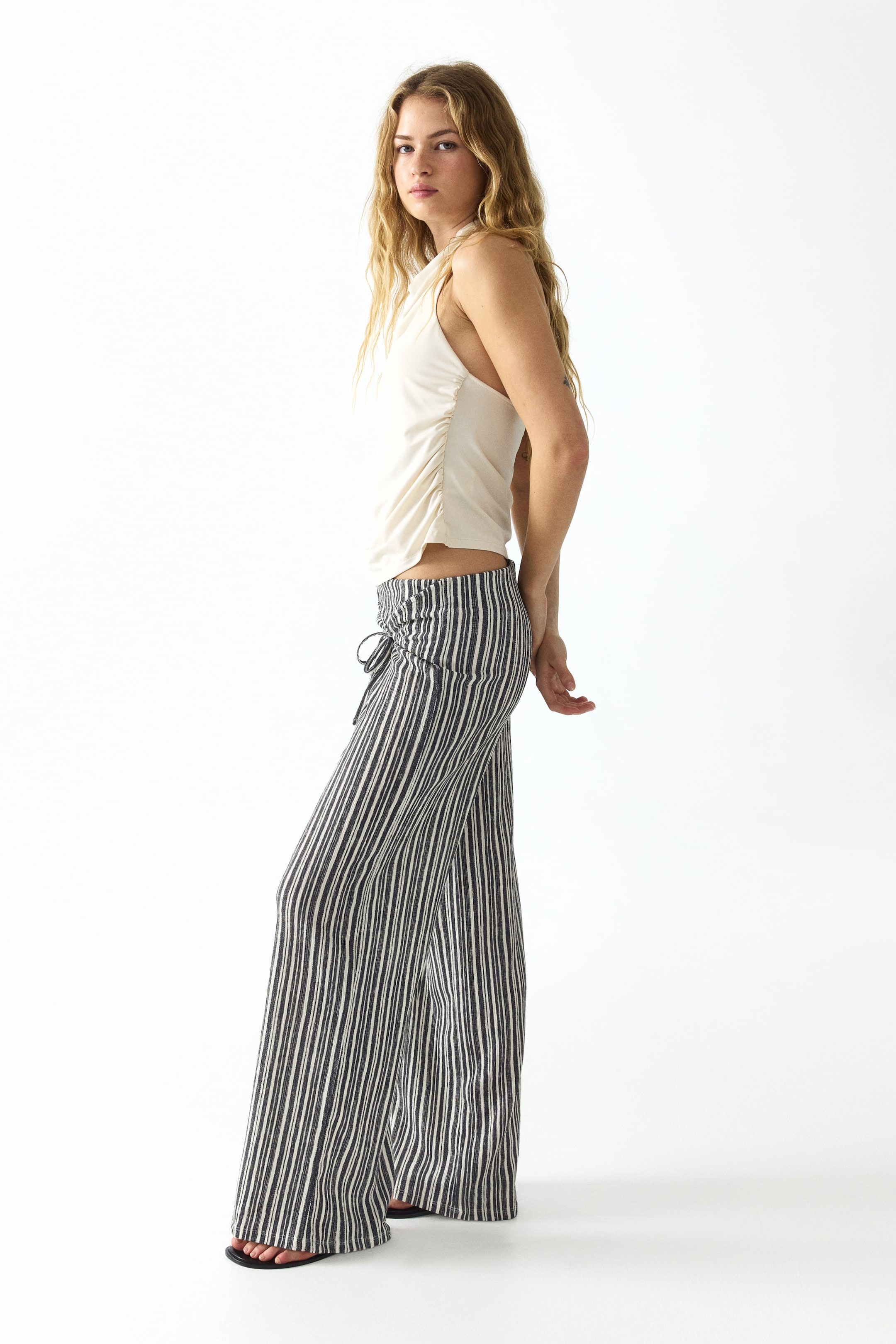 Agrandir l'image: Le mannequin porte un pantalon en jersey texturé avec des rayures verticales noires et blanches. Une large ceinture à nouer se trouve à l'avant de la taille basse, et les jambes droites tombent avec élégance.