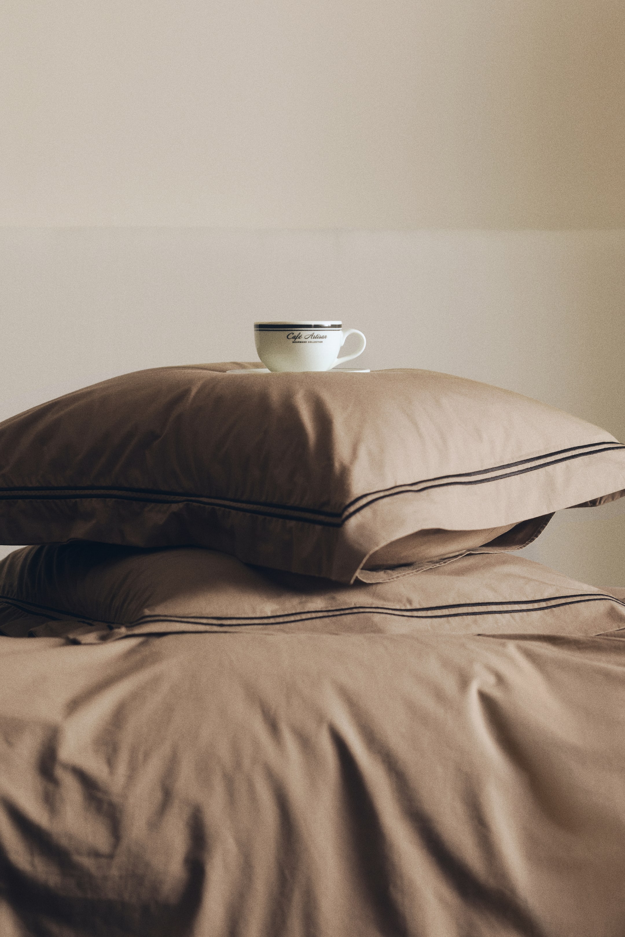 View larger image: Showing a taupe-brown cotton quilt cover set featuring two stacked pillowcases with dark, double-embroidered parallel lines along the edges. A white ceramic coffee cup and saucer are resting on top of the upper pillow.