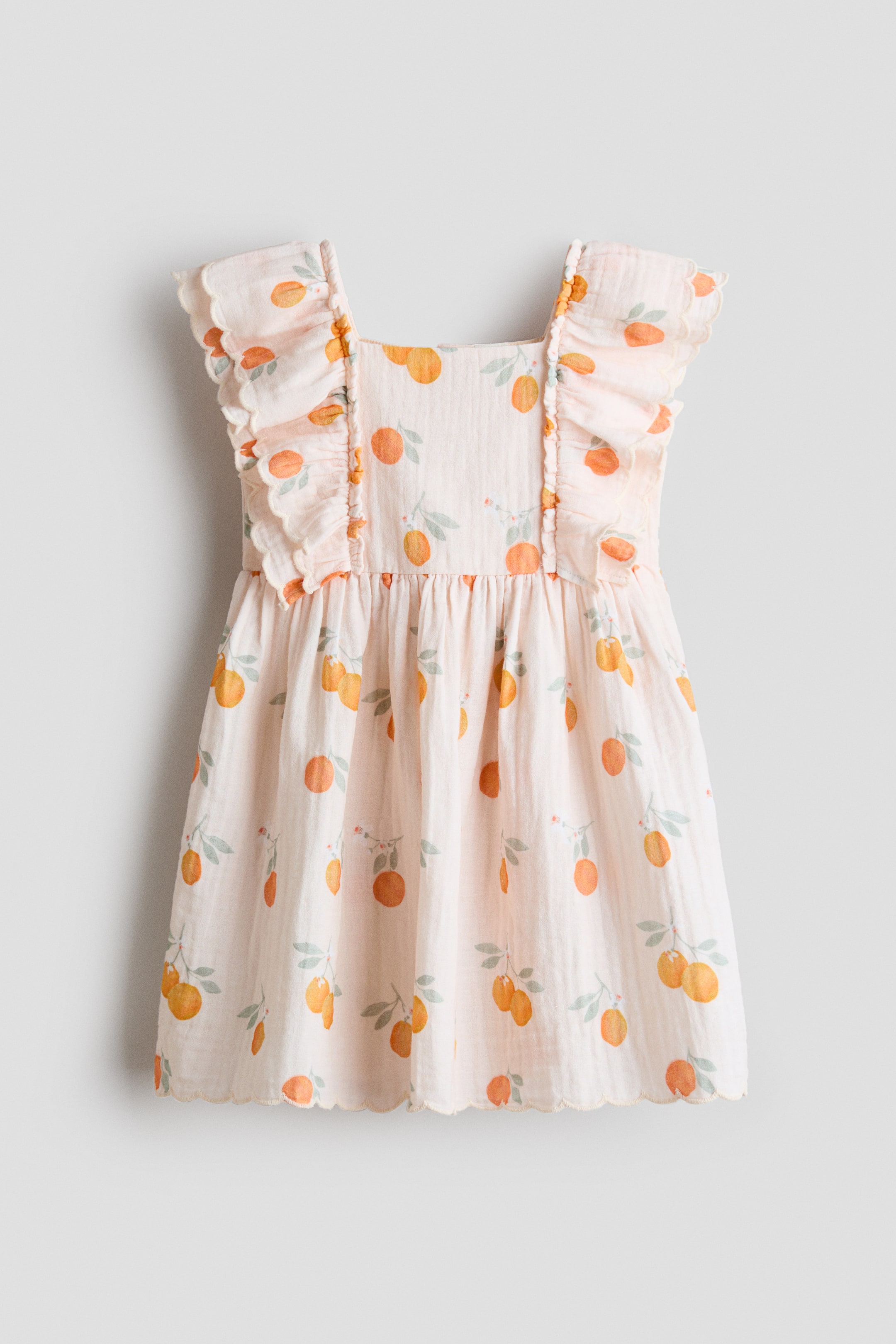 Visa större bild: H&M Adorables ärmlösa klänning visas framifrån och visar ett mjukt vävt tyg med ett mönster av orange frukt och gröna blad. Den har en fyrkantig halsringning, ett smalt ok med dubbla lager volanger och vågiga kanter som sträcker sig över axlarna, med rynkor som bildas i midjan och leder till en utsvängd kjol med vågig fåll.