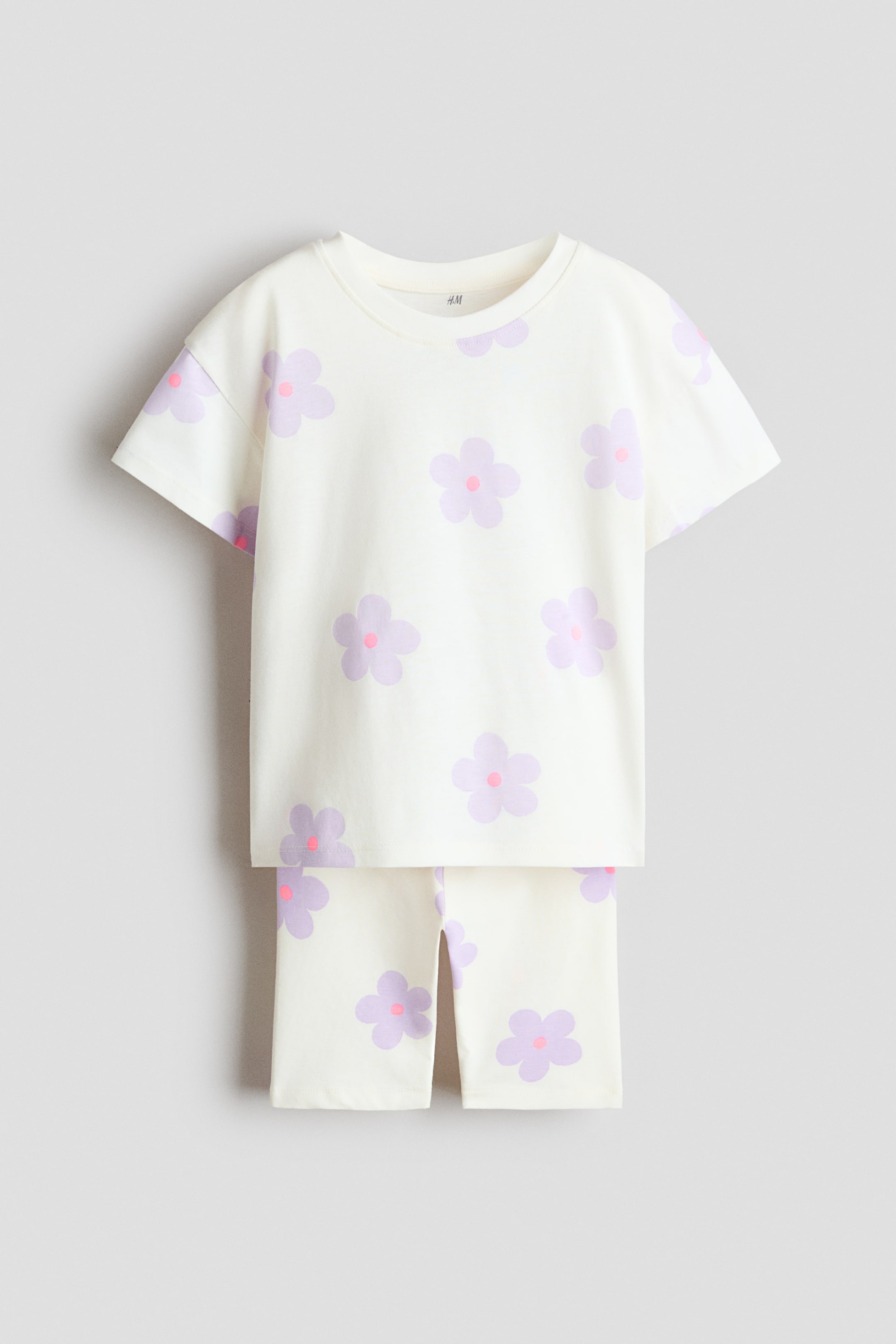 View larger image: A front-facing set features a cream T-shirt and matching cycling shorts, both showcasing a light purple floral print with pink centers. The T-shirt has a ribbed neckline and dropped shoulders, while the cycling shorts have a smooth, elastic waistband.