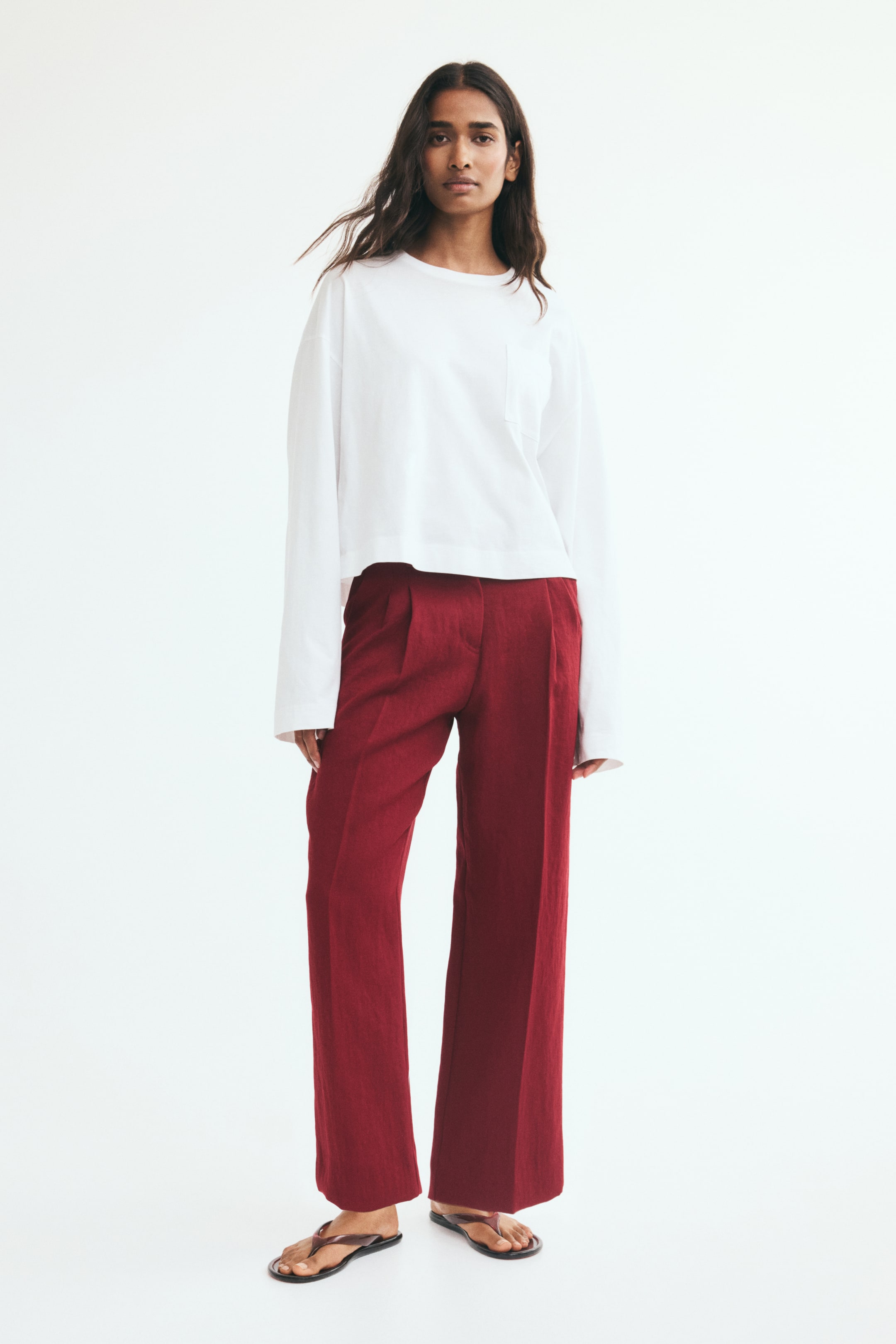 View larger image: Dark red, loose-fit tailored trousers with straight legs, front pleats, and pressed creases are worn by the model. The ankle-length trousers are styled with a long-sleeved white top and flat sandals.